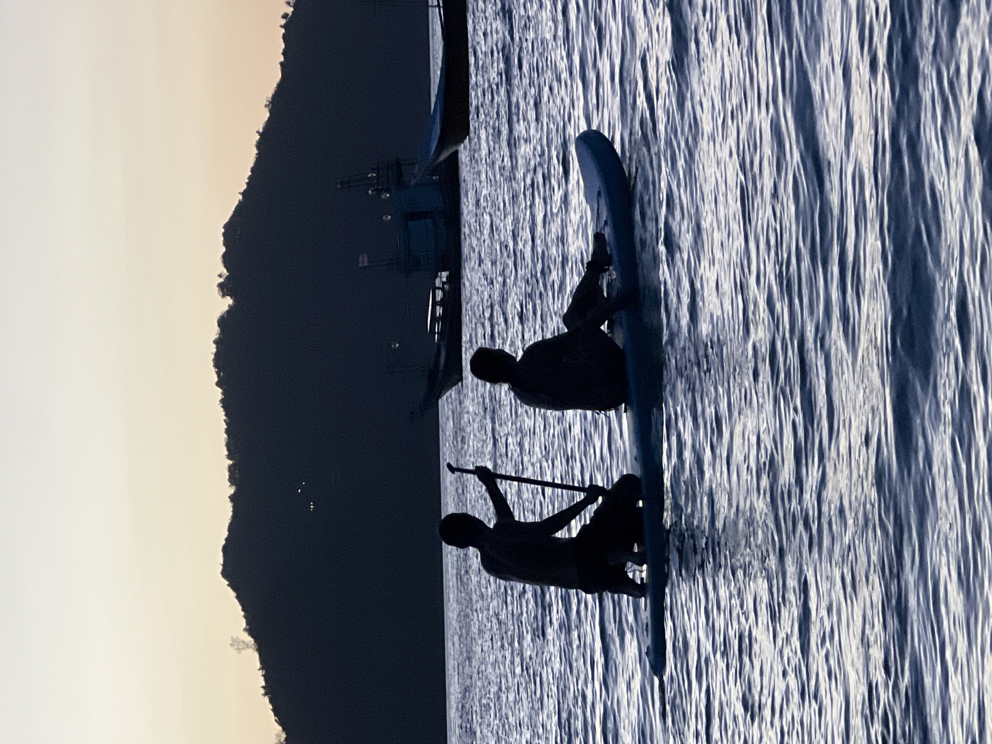 This media file depicts a serene scene of two individuals paddleboarding on calm water, likely at dusk or dawn, given the soft, diffused light. The silhouette of a mountainous coastline dominates the left side of the frame, with a faint glow on the horizon suggesting either sunrise or sunset. In the background, a boat or ferry is partially visible against the dark landmass, hinting at a coastal or island setting.
The primary subjects are two people on separate paddleboards. The person in the foreground is seated on their board, holding a paddle that rests on the water. Their posture suggests a moment of rest or observation. Further back, another individual is also on a paddleboard, positioned on a separate board. Their silhouette is less defined, but they appear to be in motion, possibly paddling. The water itself is a prominent element, characterized by gentle ripples that catch the light, creating a textured and dynamic surface.
The overall mood is tranquil and peaceful, with the soft lighting and calm water contributing to a sense of quietude. The silhouettes emphasize the shapes of the figures and the landscape, creating a visually striking contrast between light and shadow. No text is visible in the image. The location context provided places this scene in the Ko Pha-ngan District, Thailand, a region known for its scenic islands and coastal beauty, which aligns with the visual elements in the image.