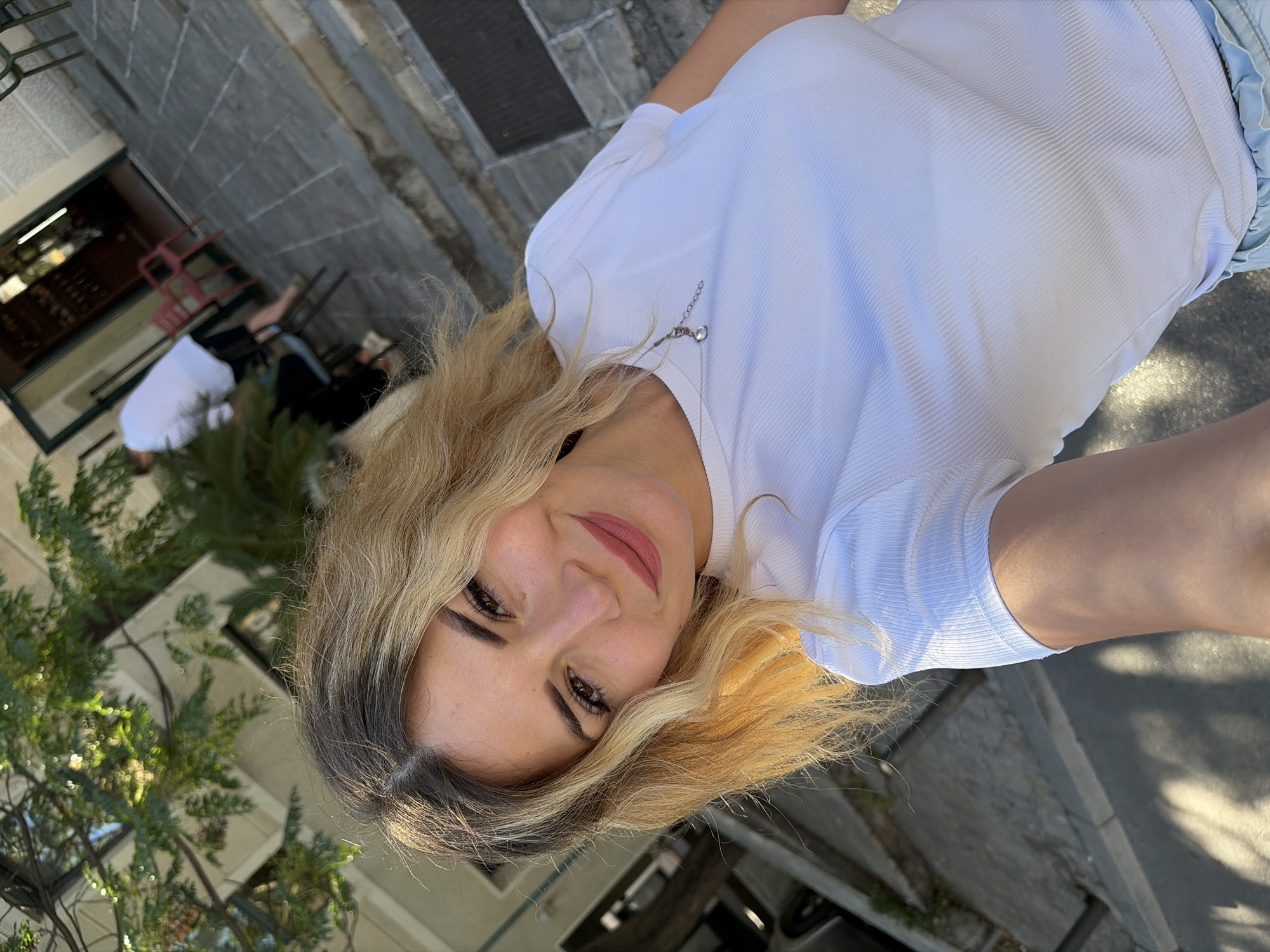 A woman with blonde hair and light skin is taking a selfie outdoors. She is wearing a white t-shirt with a delicate silver necklace. The background suggests a European city with cobblestone paving and buildings with arched doorways. In the blurred background, a person in a white shirt and dark shorts is visible, and a red chair is placed outside a doorway. The scene appears to be a street or plaza in Budva, Montenegro, as indicated by the architectural style and paving.