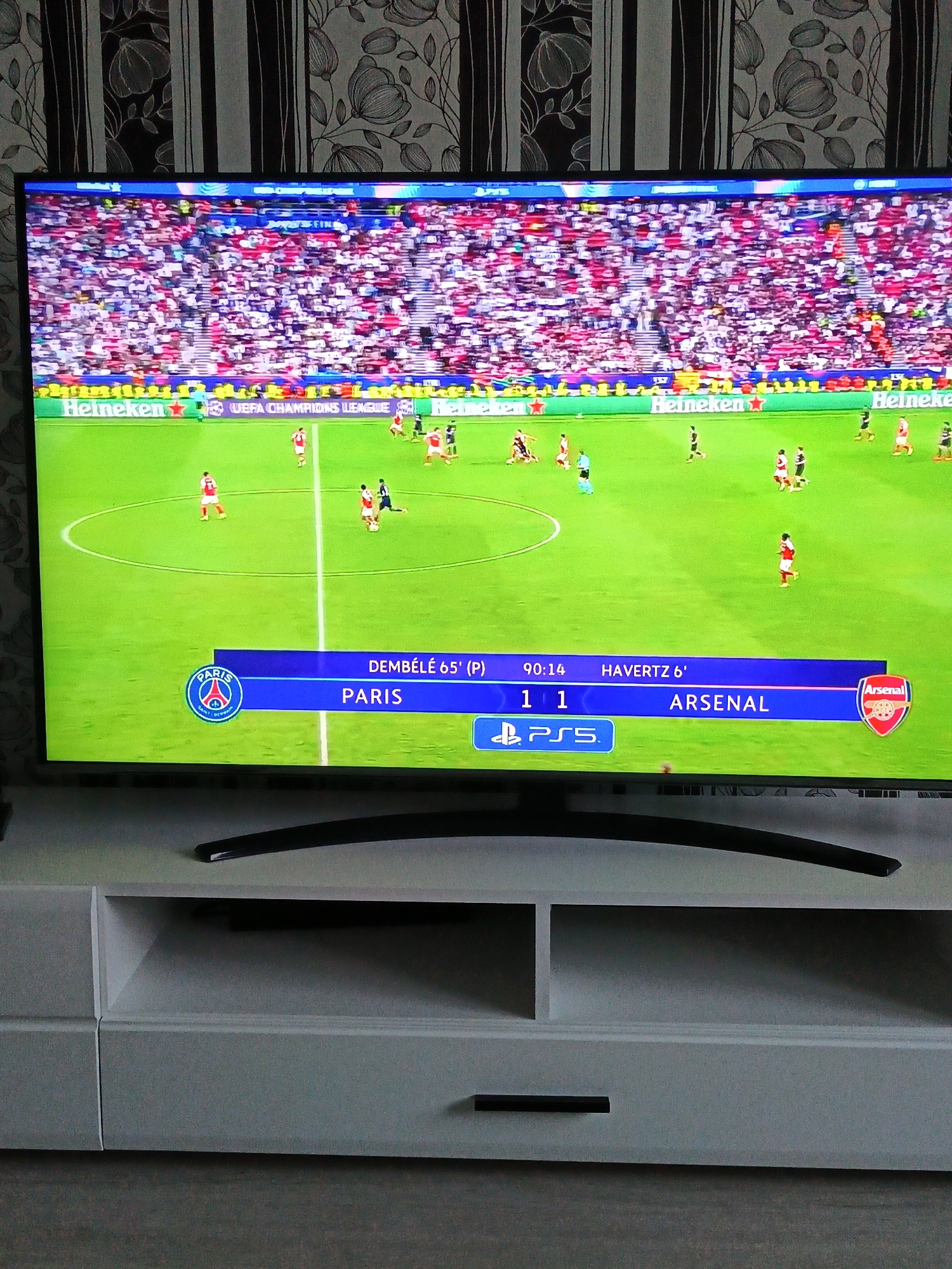 The image shows a television screen displaying a soccer game between Paris and Arsenal. The score is 1-1 in the 90:14 minute, with Dembélé scoring for Paris in the 65th minute and Havertz scoring for Arsenal in the 6th minute. The background shows a crowd of spectators in the stadium. The television is placed on a white TV stand with drawers and cabinets. The background behind the TV is a wall with a striped floral pattern.
There is no indication the image was taken in Парэчча, Belarus.