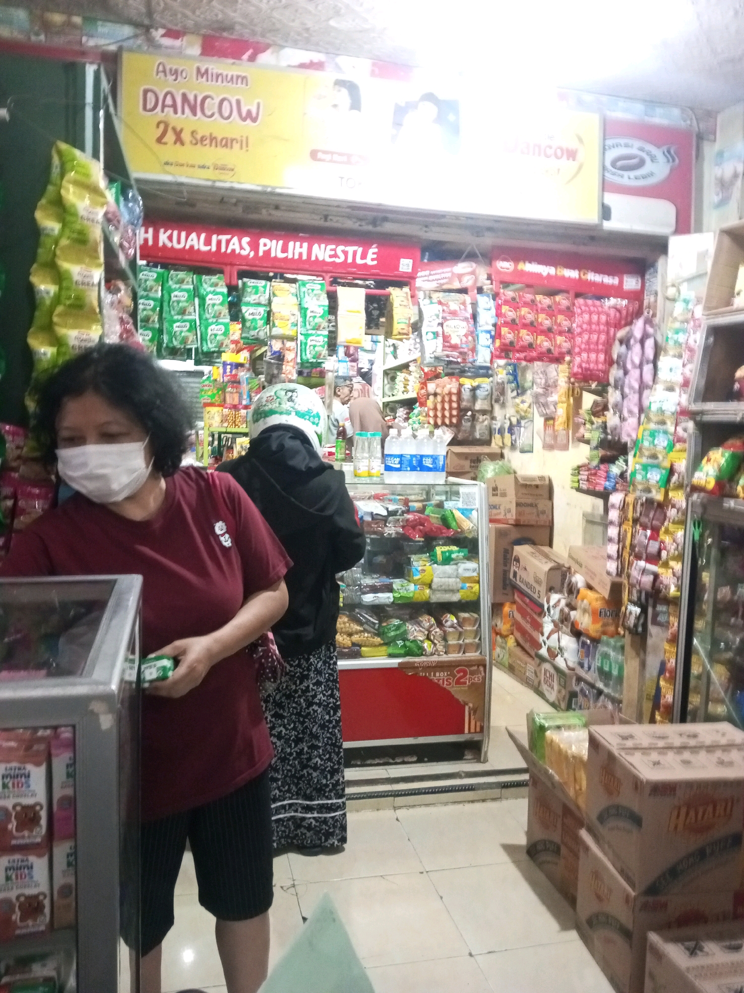 The image depicts the interior of a small convenience store, likely a warung, in Malang, Indonesia. A woman wearing a maroon t-shirt and dark striped shorts, with a white surgical mask covering her face, stands in the foreground to the left, holding a small green package. Behind her, another person, partially obscured by a black hooded jacket and a white helmet adorned with a cartoonish design, is visible. The store is densely stocked with various products, including bags of snacks, canned goods, bottled drinks, and boxed items stacked on shelves and on the floor. Prominent signage includes a yellow banner advertising "Dancow 2x Sehari!" and another red banner with "H KUALITAS, PILIH NESTLE." Boxes of "HATARU" brand products are visible on the right side. The overall environment suggests a typical, bustling local shop.