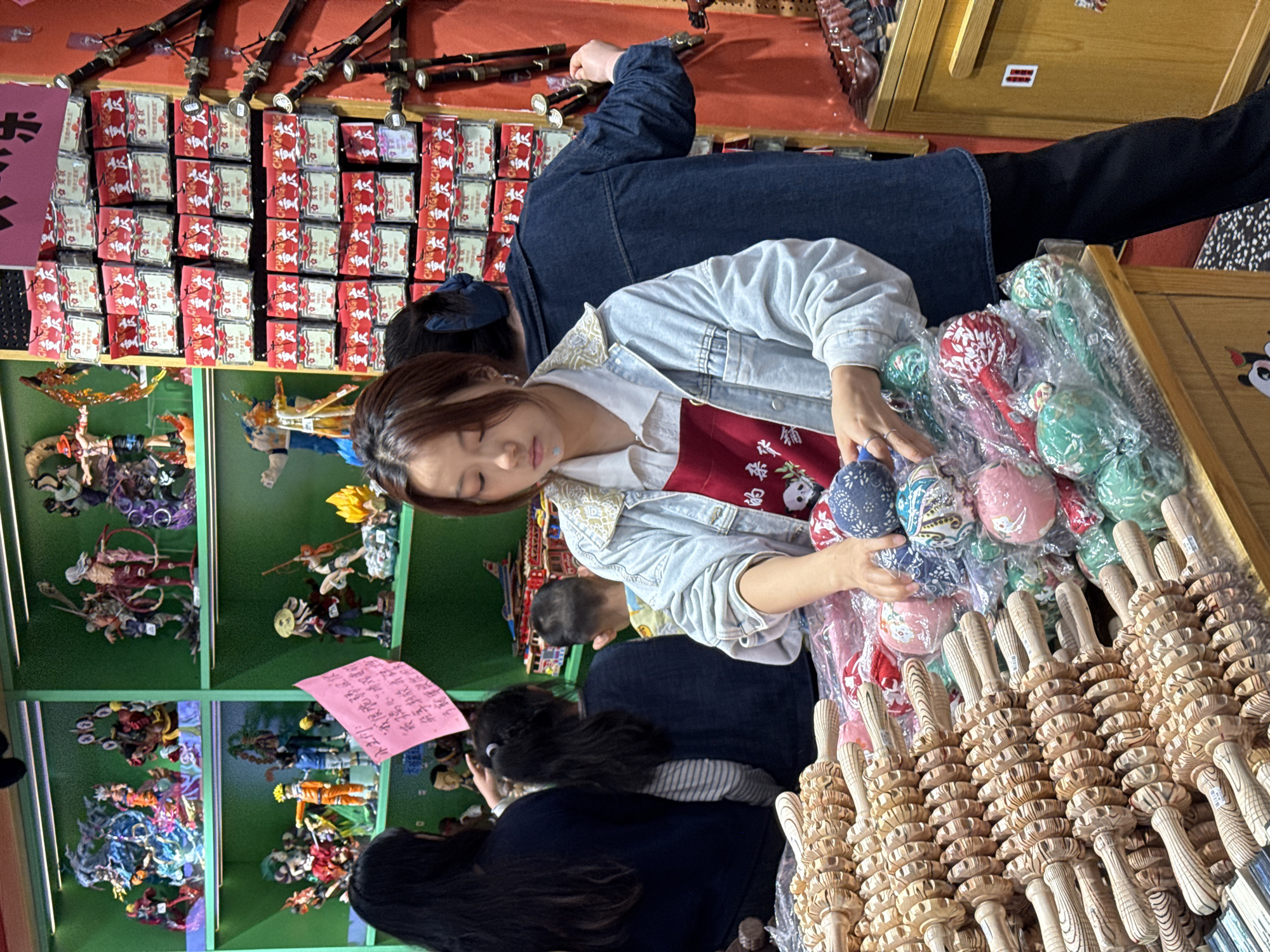 This indoor image depicts a bustling retail environment within the Shapingba District of China. The focal point is a woman wearing a light-colored jacket and a red top, examining a collection of multicolored, patterned balls encased in clear plastic. In the foreground, a large assortment of wooden massage rollers are arranged, their handles pointing towards the bottom of the frame.
Behind the woman, there are other customers browsing the merchandise. To the left, a brightly lit display case houses various action figures on green shelves. Above this, red packages with white motifs are neatly arranged. Further in the background, indistinct figures suggest a continued shopping environment.
The scene conveys an active commercial space, with customers engaged in selecting merchandise from diverse product categories.