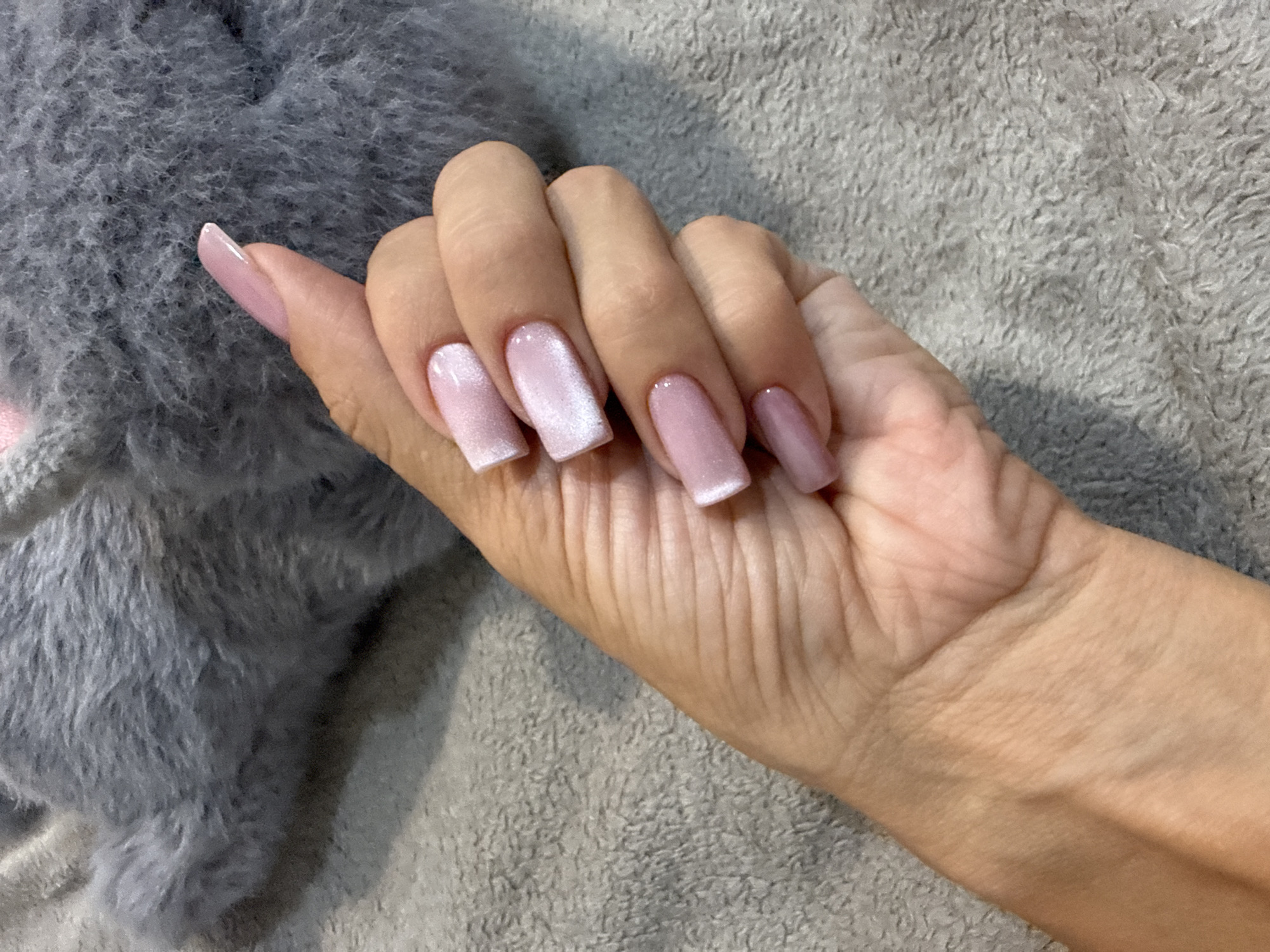 A person's hand with light pink, square-shaped acrylic nails is resting on a textured gray surface. The nails have a subtle shimmer, and the skin of the hand shows natural lines and a faint vein on the wrist. A blurry, soft gray object, possibly a plush toy or blanket, is visible in the upper portion of the image. The lighting suggests an indoor setting. The image does not provide any information related to Rusanovo, Russia.