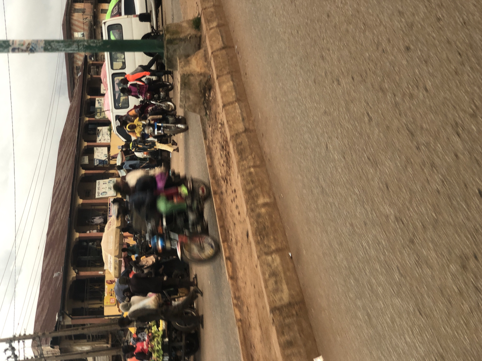 The media file depicts a busy street scene in Bukuru, Nigeria. The primary subjects are numerous motorcycles, many with riders, parked and in motion. People are visible interacting with the motorcycles and walking along the street, suggesting a marketplace or transport hub.
The scene appears to be set during the daytime, under an overcast sky, as indicated by the diffused light and lack of strong shadows. The environment shows a row of buildings with arched windows and verandas, characteristic of local architecture. A white van is also present in the background. The street is paved with asphalt and bordered by a raised concrete curb. The overall impression is one of a bustling, active urban environment.
Notable details include the various colors of clothing worn by the people, and the presence of a sign with text, though it is largely illegible due to motion blur and distance. The motion blur on the motorcycles and some of the people suggests activity and movement, possibly indicating a busy commute or commerce. No specific emotions are discernible due to the quality of the image. The location cues of the architecture and the type of vehicles strongly suggest a Nigerian setting, consistent with the provided location context.