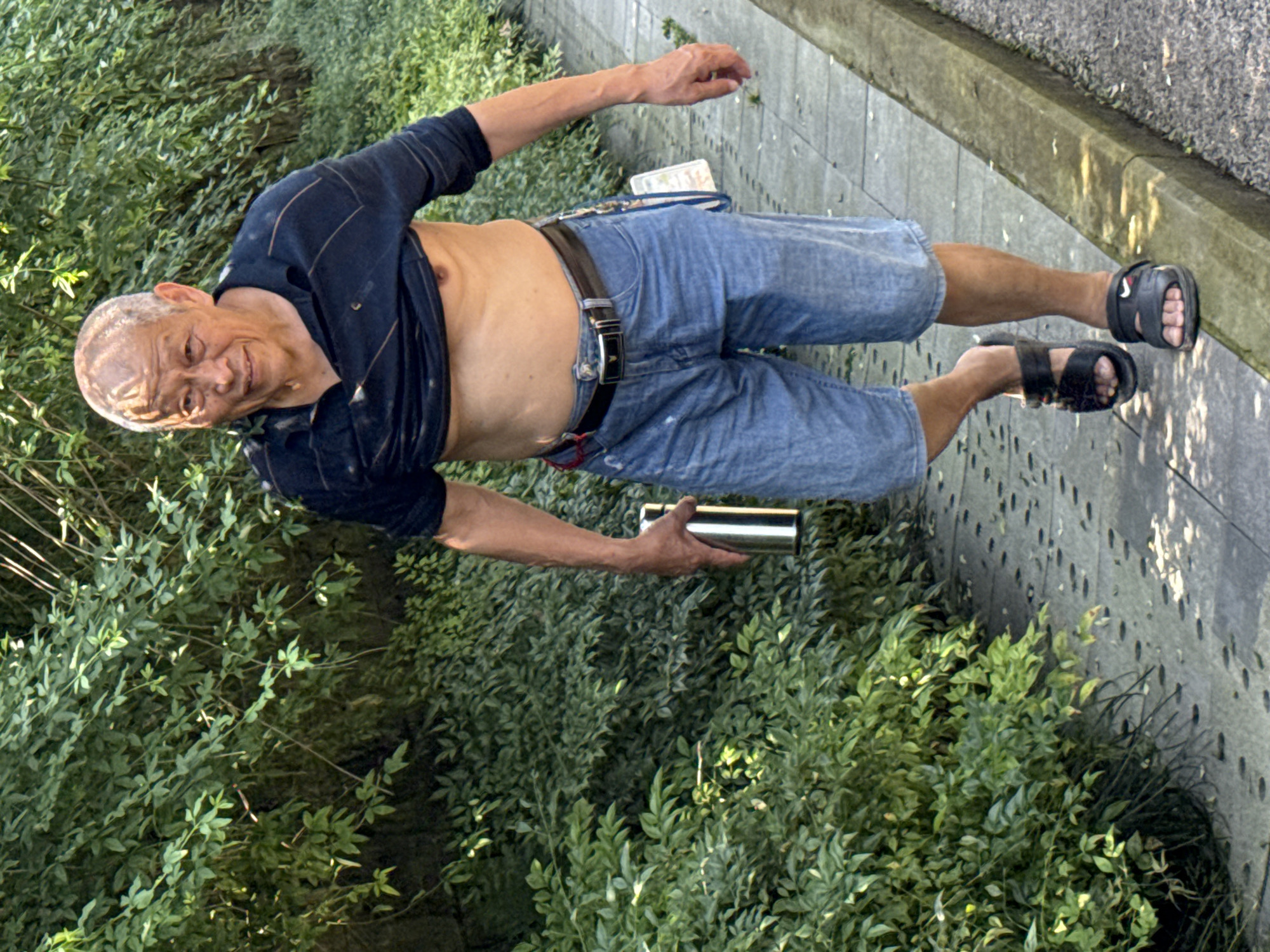 An older man stands outdoors, captured in a slightly low-angle shot, looking towards the viewer with a relaxed expression and a subtle smile. He is positioned on a paved walkway, with lush green bushes and foliage to his left and behind him, and a paved street or pathway to his right.
The man is wearing light blue denim shorts or cutoff jeans, a dark blue long-sleeved shirt that is pulled up, exposing his bare midriff and chest, and black sandals. A brown leather belt with a metal buckle is visible around his waist. In his left hand, he holds a silver, metallic cylindrical container, resembling a thermos or water bottle. His right arm is bent, with his hand resting near his hip.
His skin appears tanned, and his head shows some patterns on the scalp, possibly from light reflecting off short hair or a hair treatment. On the strap of his right sandal, a white Nike logo is visible.
The setting appears to be a sunny day, with bright ambient light and some shadows cast on the paved ground, suggesting either morning or late afternoon. The overall scene conveys a casual, outdoor moment in what appears to be a public area or park in Banan, China.