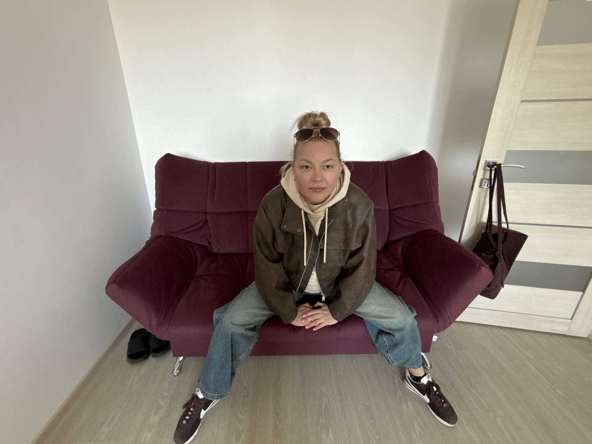 A person is seated on a burgundy sofa in an indoor setting. The person is wearing blue jeans, brown sneakers with white accents, a beige hooded sweatshirt, and a brown jacket. Sunglasses are perched atop their head. Black slides are visible next to the sofa. A brown purse hangs from a door to the right of the sofa. The walls are white. The floor is light-colored wood or laminate. The location cannot be determined to be in Izhevsk, Russia from this image alone.