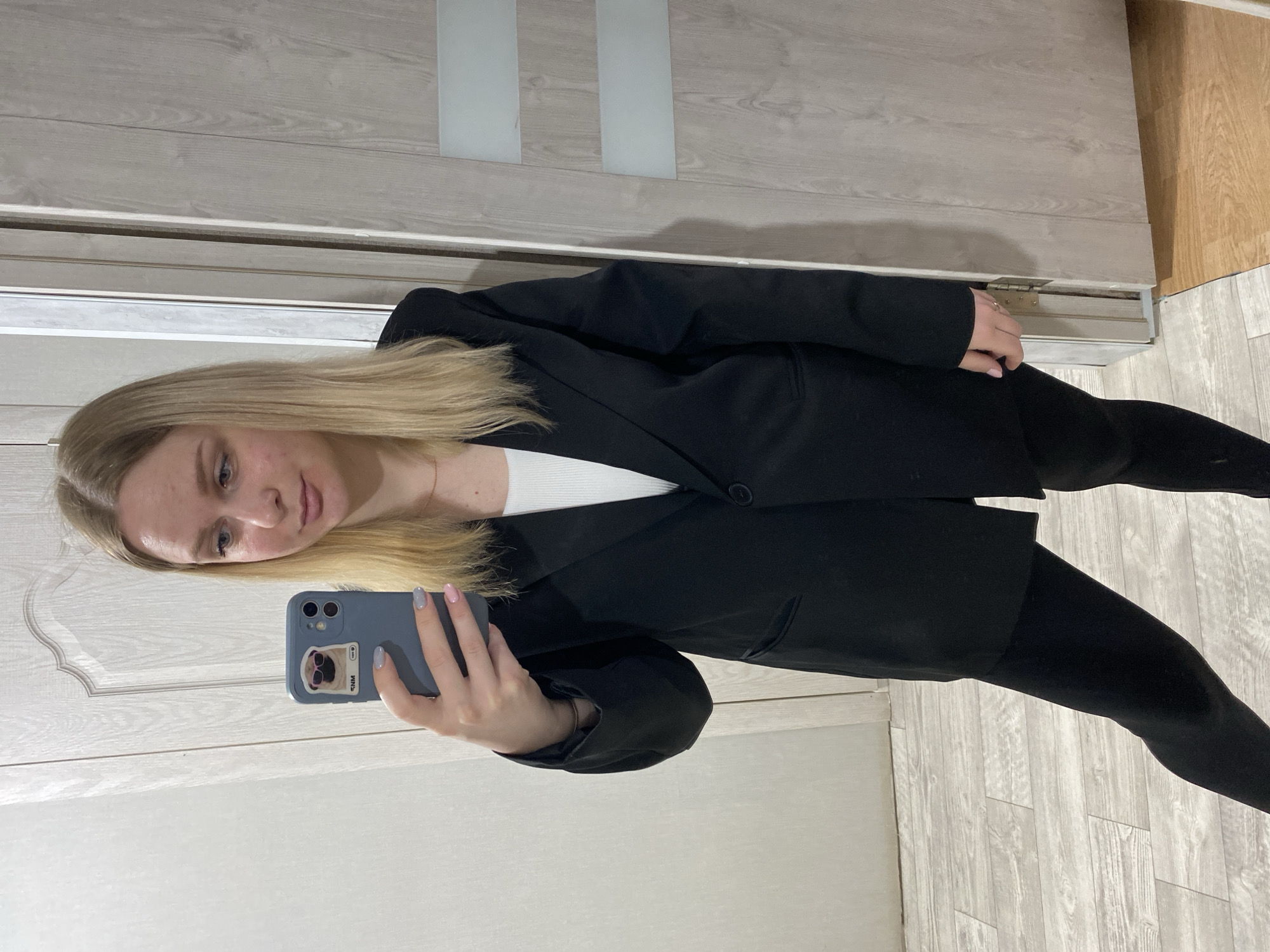 In an indoor setting within Unknown, Unknown, a blonde-haired female is captured in a mirror selfie, standing mostly upright. She has fair skin with visible facial blemishes and partially visible blue eyes. Her hair is straight, light blonde, and parted on her left side. She is wearing a black single-button blazer over a white ribbed tank top and black leggings. A thin gold-colored necklace is visible around her neck. On her right wrist, a thin gold-colored bracelet is present, along with a silver-colored ring on her right ring finger. Her left hand holds a blue/grey smartphone with a dual camera setup, featuring a sticker of a pug dog wearing red sunglasses and the text "SNM". Her nails are manicured with light grey polish on the thumb and index finger, and light pink polish on the middle and ring fingers. The background consists of light wood-grained doors; the left door has a decorative carved panel, while the right door features two horizontal frosted glass panels. The floor is composed of light grey wood-look tiles transitioning to a warmer brown wood-look flooring on the right, with a door hinge visible near the bottom right.