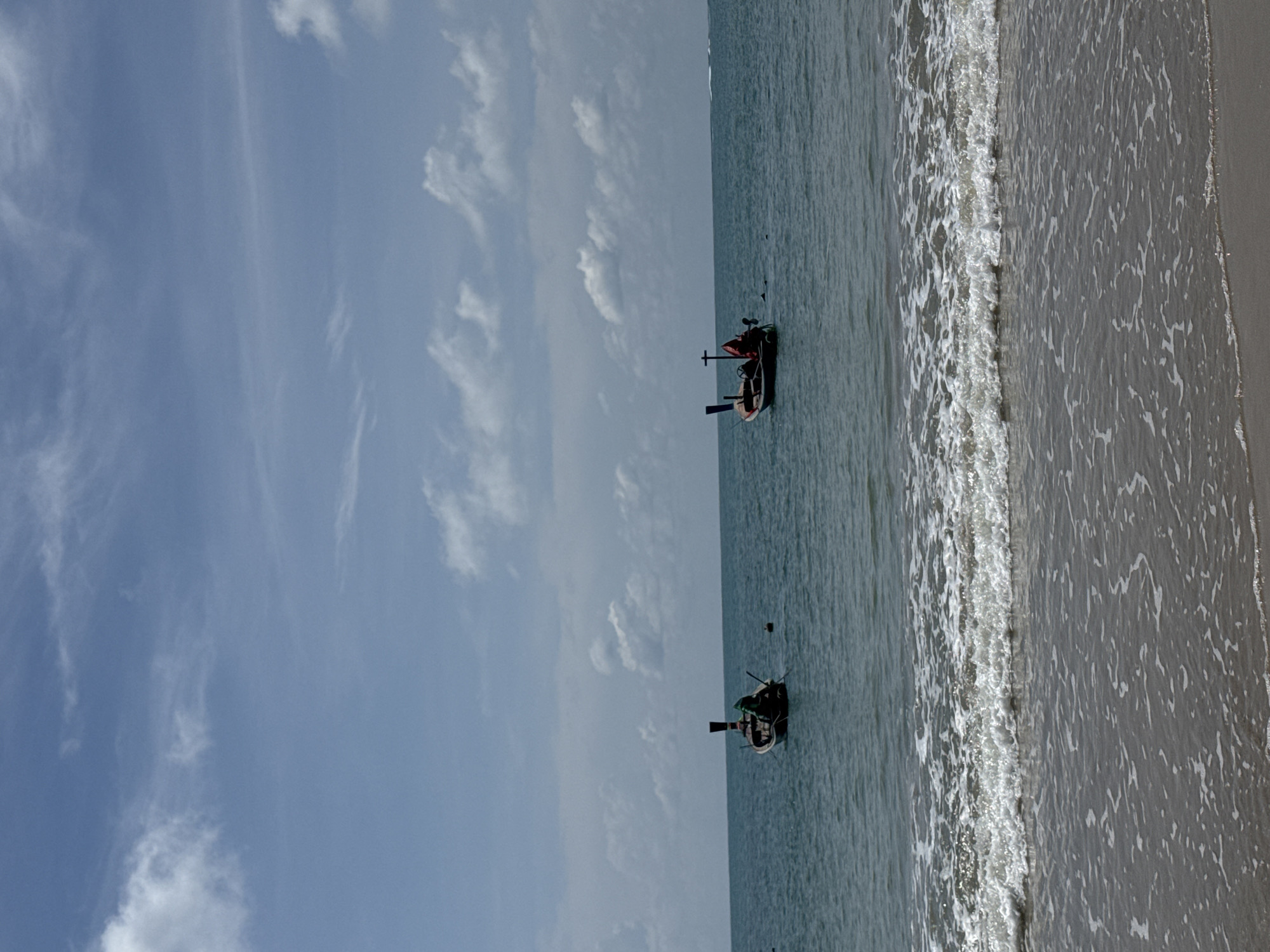 This image captures a serene coastal scene with two long-tail boats floating in the turquoise waters of Sakhu, Thailand. The boats are positioned at a distance from the shore, where gentle waves with white foam are breaking on a sandy beach. The sky above is a clear blue, adorned with scattered white clouds, suggesting a bright and pleasant day. The composition is dominated by the sea and sky, creating a sense of openness and tranquility.