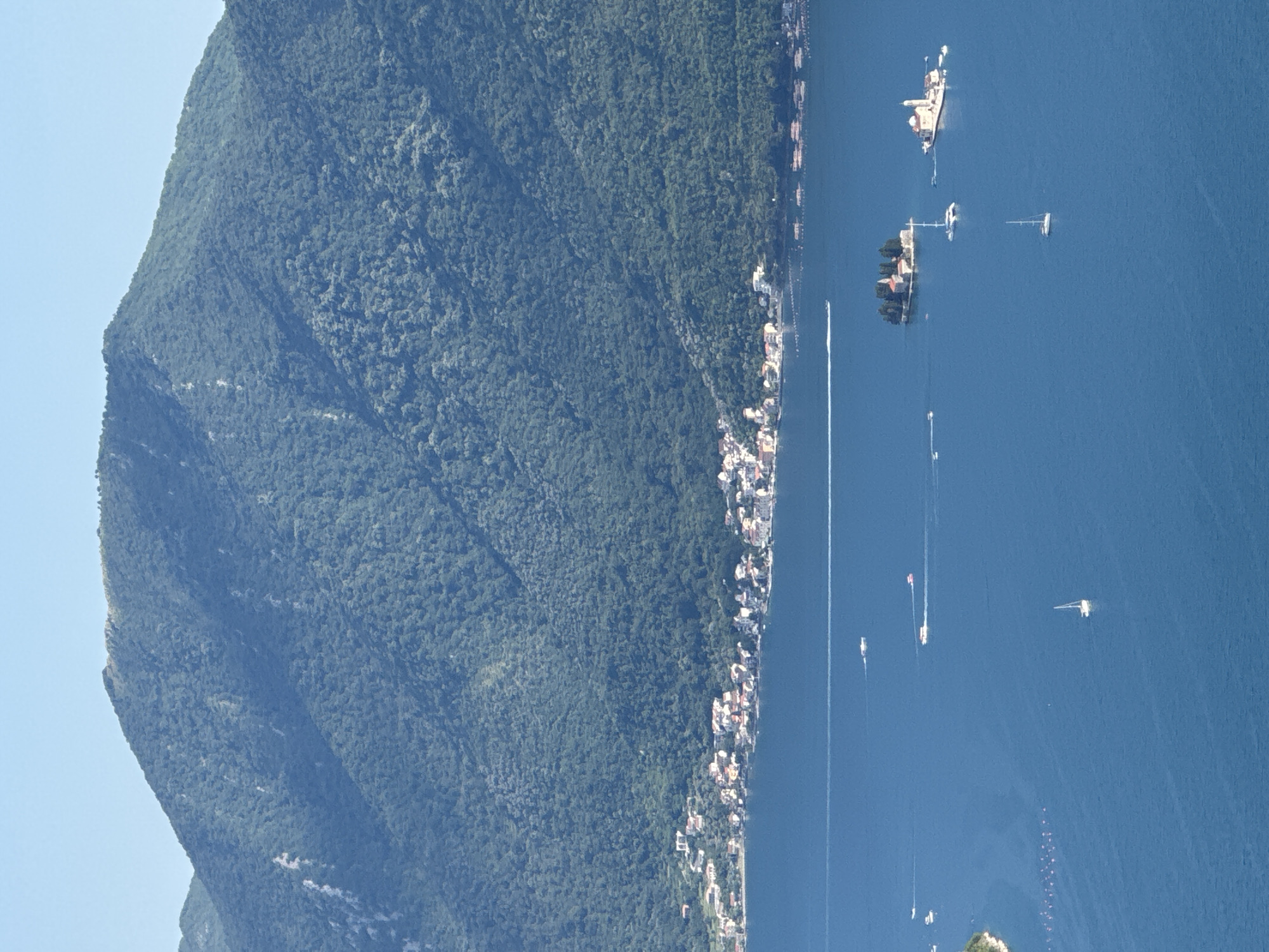 The image depicts a wide, elevated view of a coastal area in Strp, Montenegro. In the foreground, a vast expanse of deep blue water dominates the scene, with several boats scattered across its surface. One sailboat is visible, its white sail clearly defined against the blue. Further out, a few motorboats are present, leaving faint white wakes.
In the middle ground, two small, verdant islands are prominent. The island to the left features a cluster of buildings, appearing to be a small village or settlement. The island to the right is topped with a distinct white church, characteristic of the region.
The background is dominated by a large, imposing mountain covered in dense green vegetation. The slopes of the mountain are steep and textured, with visible patches of lighter rock or soil. Along the base of the mountain, bordering the water, is a picturesque coastal town with buildings featuring red-tiled roofs and white walls, interspersed with greenery. The sky above is a clear, pale blue. The overall impression is one of a serene, sunlit day in a beautiful Mediterranean coastal landscape.