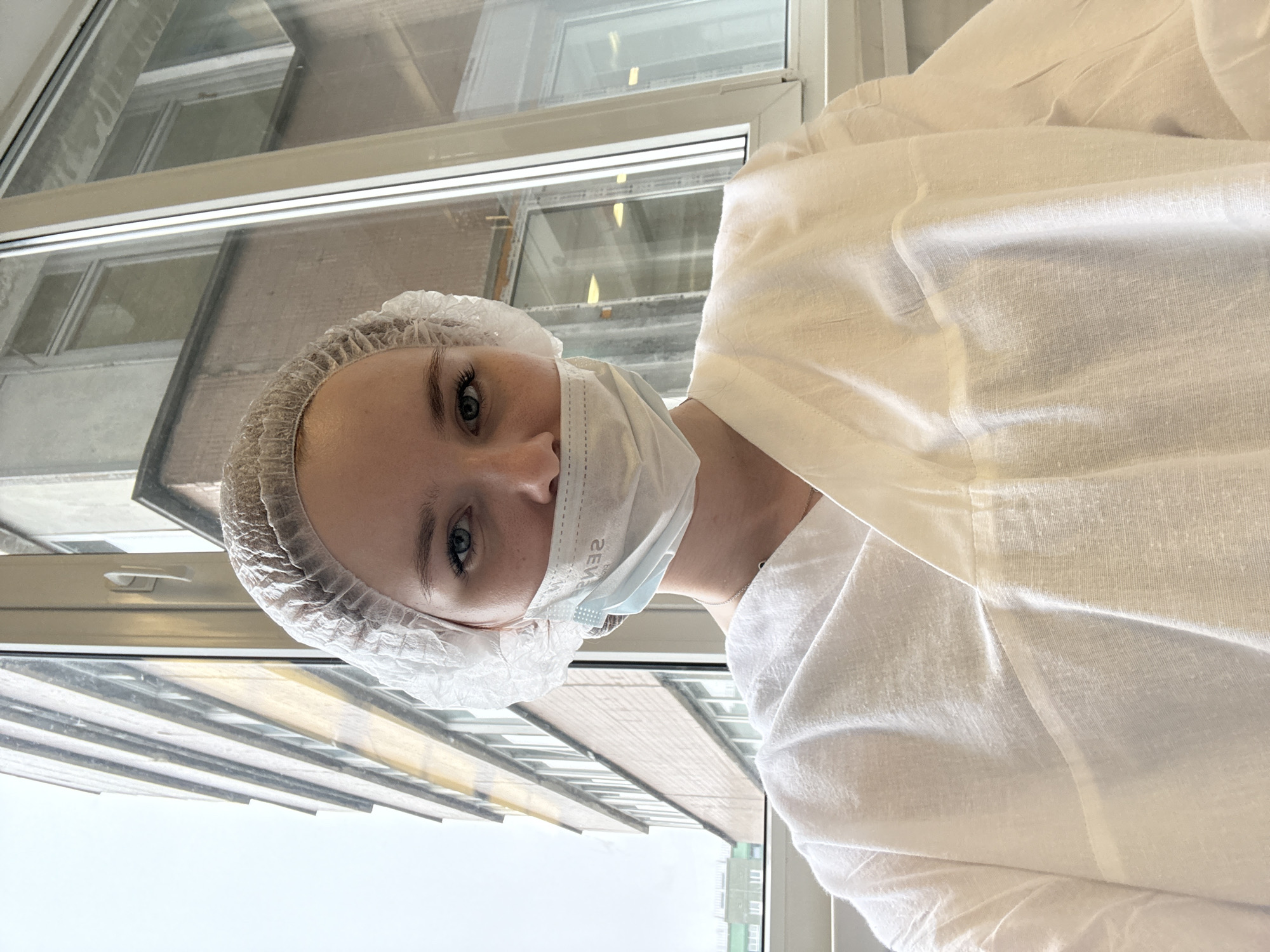 A young woman wearing a light blue medical mask, a white hair net, and a white lab coat stands indoors, facing the camera with a slight tilt of her head. Her eyes are blue. She is positioned in front of a large window. Outside the window, one can discern a building and the sky, suggesting a daytime setting. Based on the style and architecture visible through the window, the location appears to be Saint Petersburg, Russia.