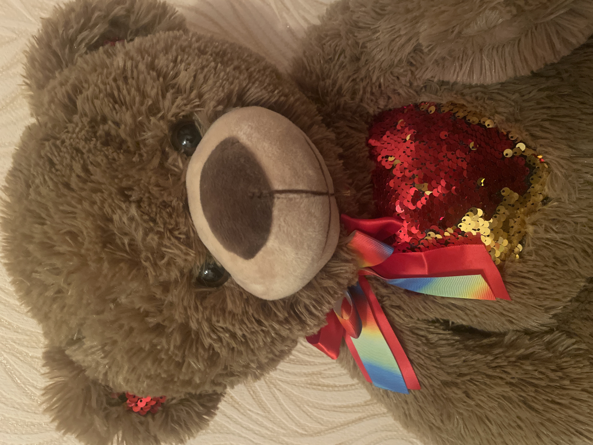 A close-up, eye-level shot shows a fluffy brown teddy bear against a textured off-white background. The bear has dark eyes, a dark brown snout, and is adorned with a bright red bow featuring a rainbow-colored ribbon. On its chest, a heart shape is outlined with red and gold sequins. The bear's fur is long and shaggy, and its ears have small red sequin accents. The overall impression is of a soft, comforting toy.