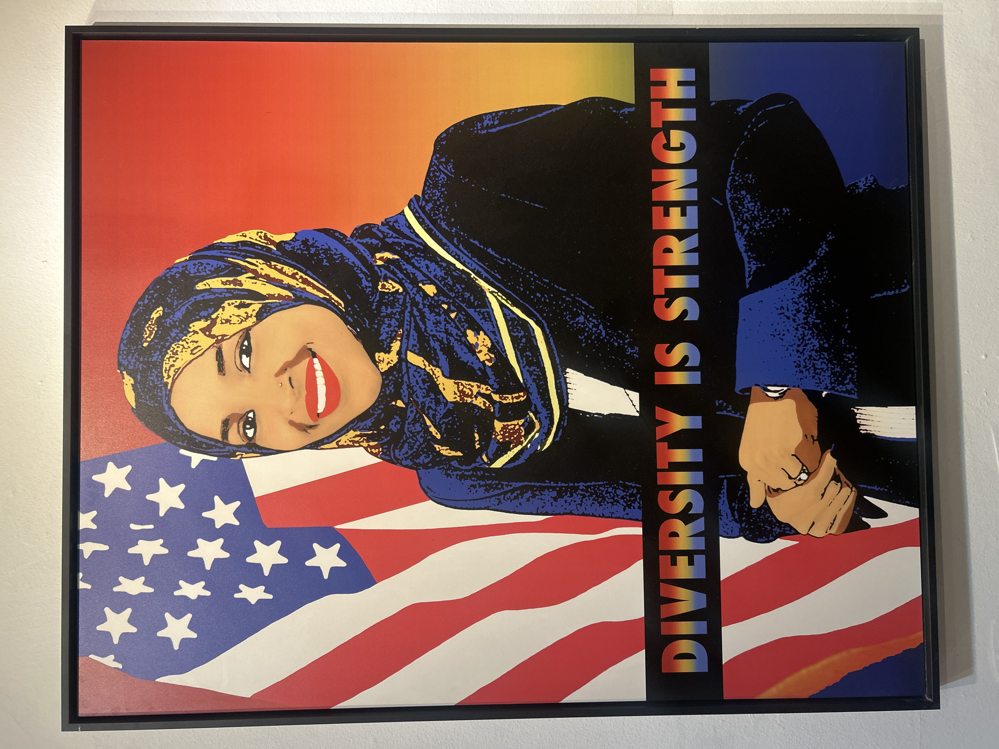 The image shows a framed poster on a white wall. The poster features a portrait of a woman wearing a hijab, smiling. The background is a gradient of red to blue, with a portion of the American flag at the bottom. The text "DIVERSITY IS STRENGTH" is printed vertically in a rainbow color scheme along the right side of the poster. The image provides no location information other than the city.