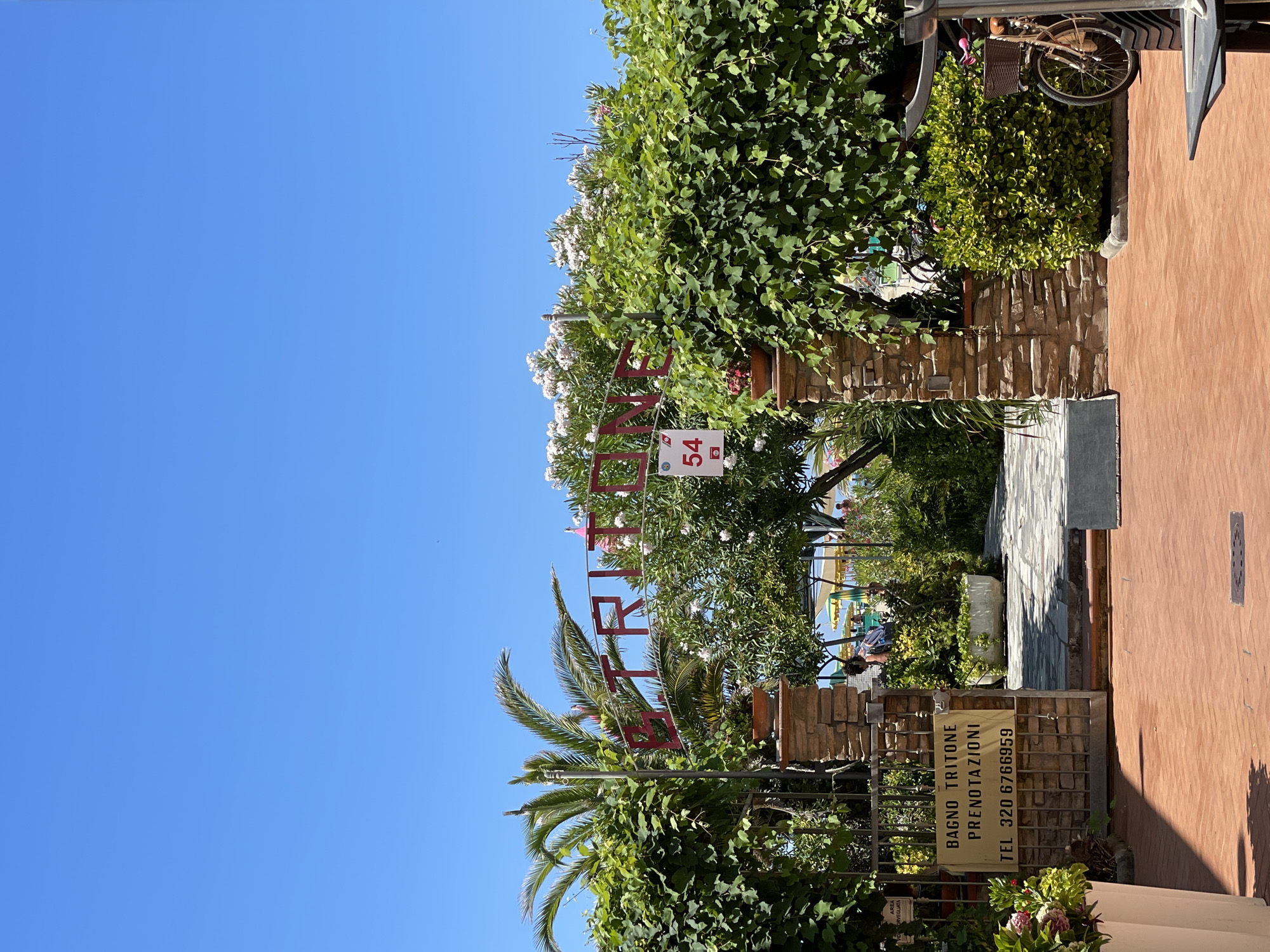 The image shows the entrance to "Bagno Tritone," a beach establishment in Viareggio, Italy. A red sign above the entrance reads "TRITONE." A smaller sign on the gate lists "BAGNO TRITONE PRENOTAZIONI" and a phone number "TEL 320 6766959." The entrance is flanked by stone pillars and abundant green foliage, with a palm tree to the left. Beyond the entrance, a pathway leads towards a beach with yellow umbrellas and people visible in the distance. A bicycle with a basket is parked to the right of the entrance. The scene is bathed in bright sunlight under a clear blue sky.