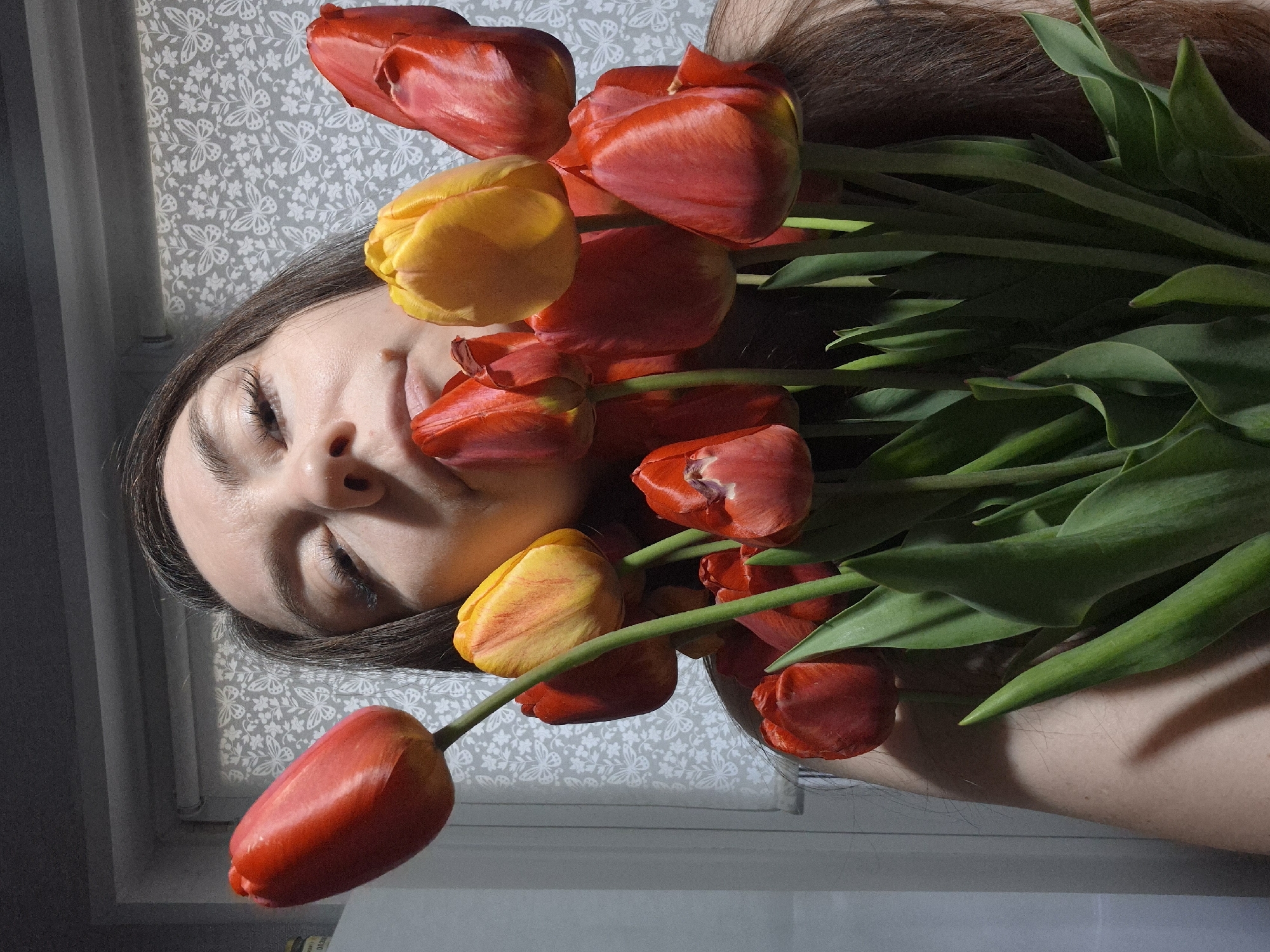 A woman with long brown hair lies on a surface patterned with small white butterflies, surrounded by red and yellow tulips. The woman's eyes are closed, and her face is partially obscured by the flowers. The scene is lit by natural light, creating shadows on the woman's face and the surrounding objects. The location is unidentified.