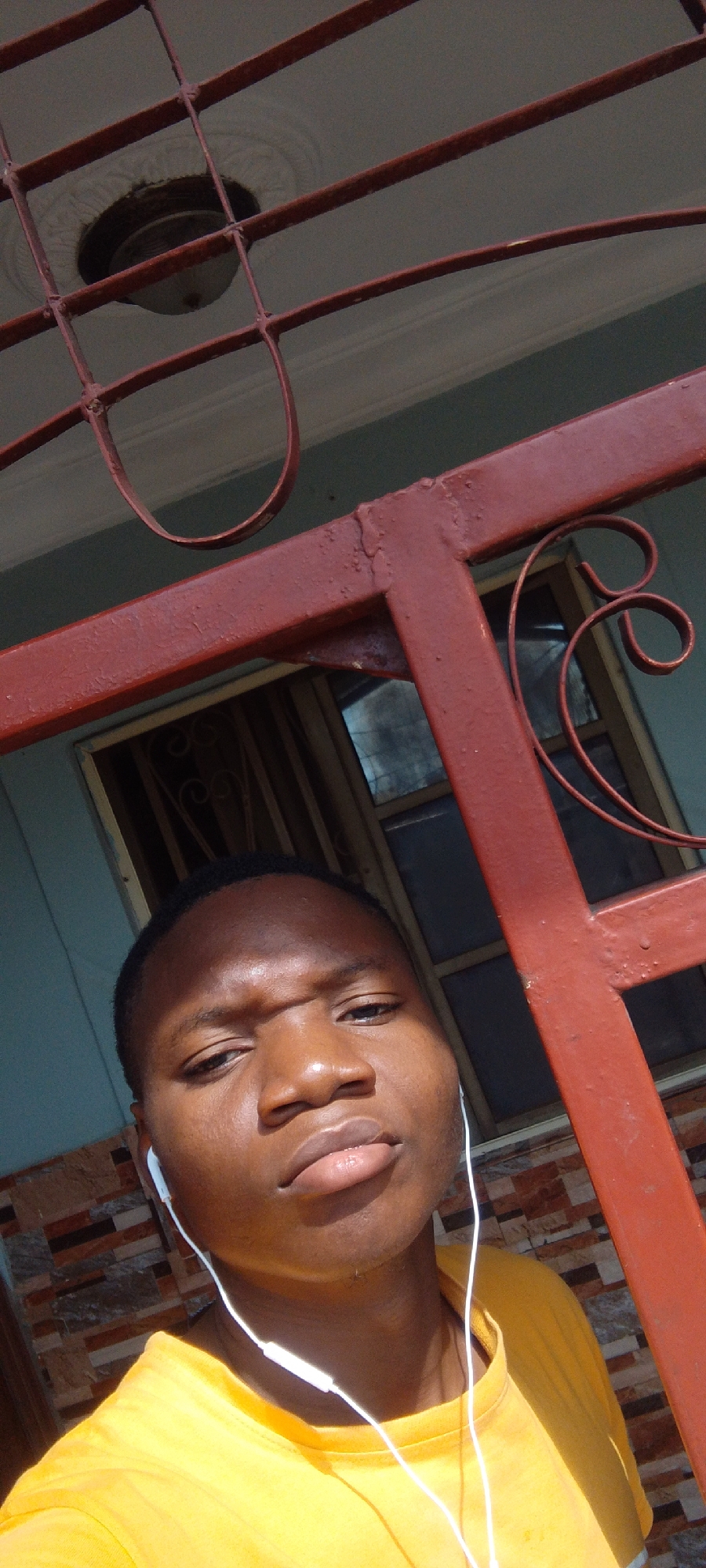 A young black male, wearing a yellow t-shirt and white earphones, is pictured from the chest up, facing the camera. He is standing outdoors, in front of a red metal gate or fence. Behind him, a building with blue walls and a window with decorative wrought iron bars is visible. The lighting suggests it is daytime, with sunlight casting shadows on the man's face and body. The background also shows a ceiling light fixture and part of a patterned wall. The overall scene appears to be residential.