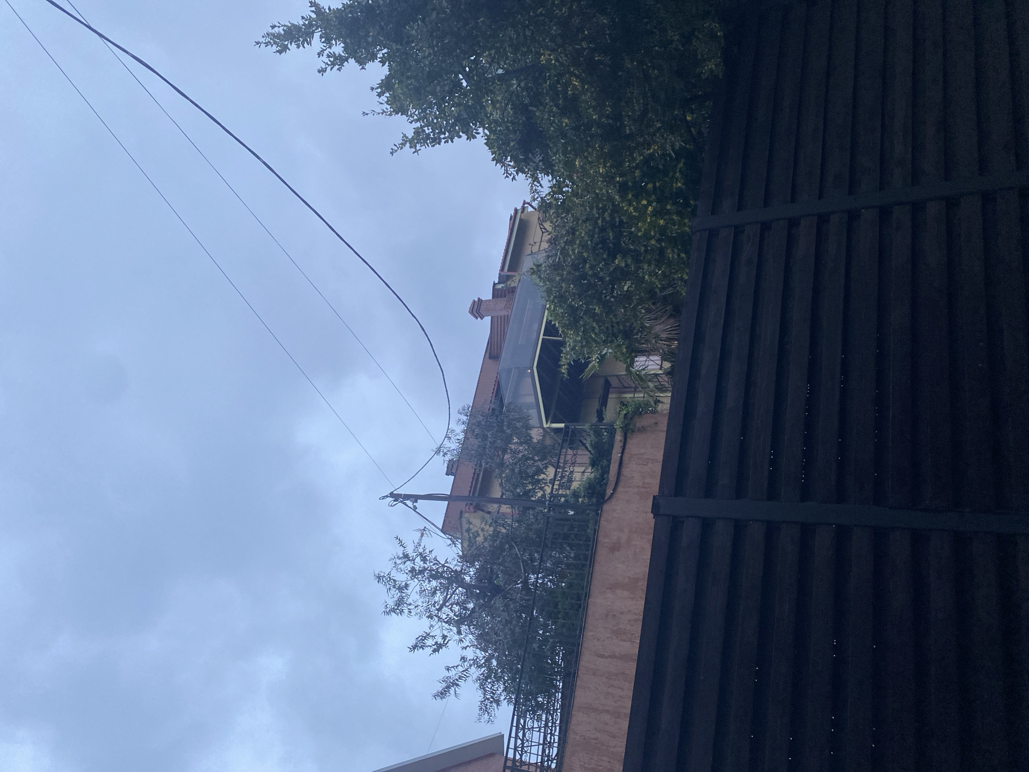 The image depicts an upward view, angled slightly, showcasing a residential scene. Dominating the right foreground is a dark, possibly black, wooden fence. Beyond the fence, a multi-story building is partially visible, featuring a salmon-colored exterior wall and balconies with metal railings. The building is adorned with greenery, including trees and plants. Power lines stretch across the upper left of the frame against a cloudy sky. The location is assumed to be in Sochi, Russia. No people are visible.