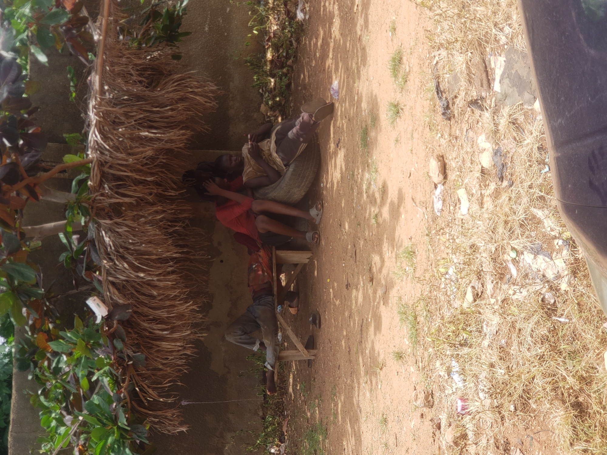 In Narayi, Nigeria, a scene unfolds under a makeshift shelter of dried palm fronds and branches, providing respite from the sun. Three individuals are visible within the shaded area. To the left, a person lies on a simple wooden bench, their legs extended and clad in blue trousers. Their upper body is partially obscured by a red and orange garment. In the center, another person, also partially clothed in red, sits with their head bent. To the right, a third individual is seated on a worn tire, holding a mobile phone. They are dressed in a light-colored sleeveless shirt and dark trousers. The ground beneath and around the shelter is composed of reddish-brown dirt, with patches of dry grass and scattered debris. A concrete wall forms the backdrop behind the shelter.