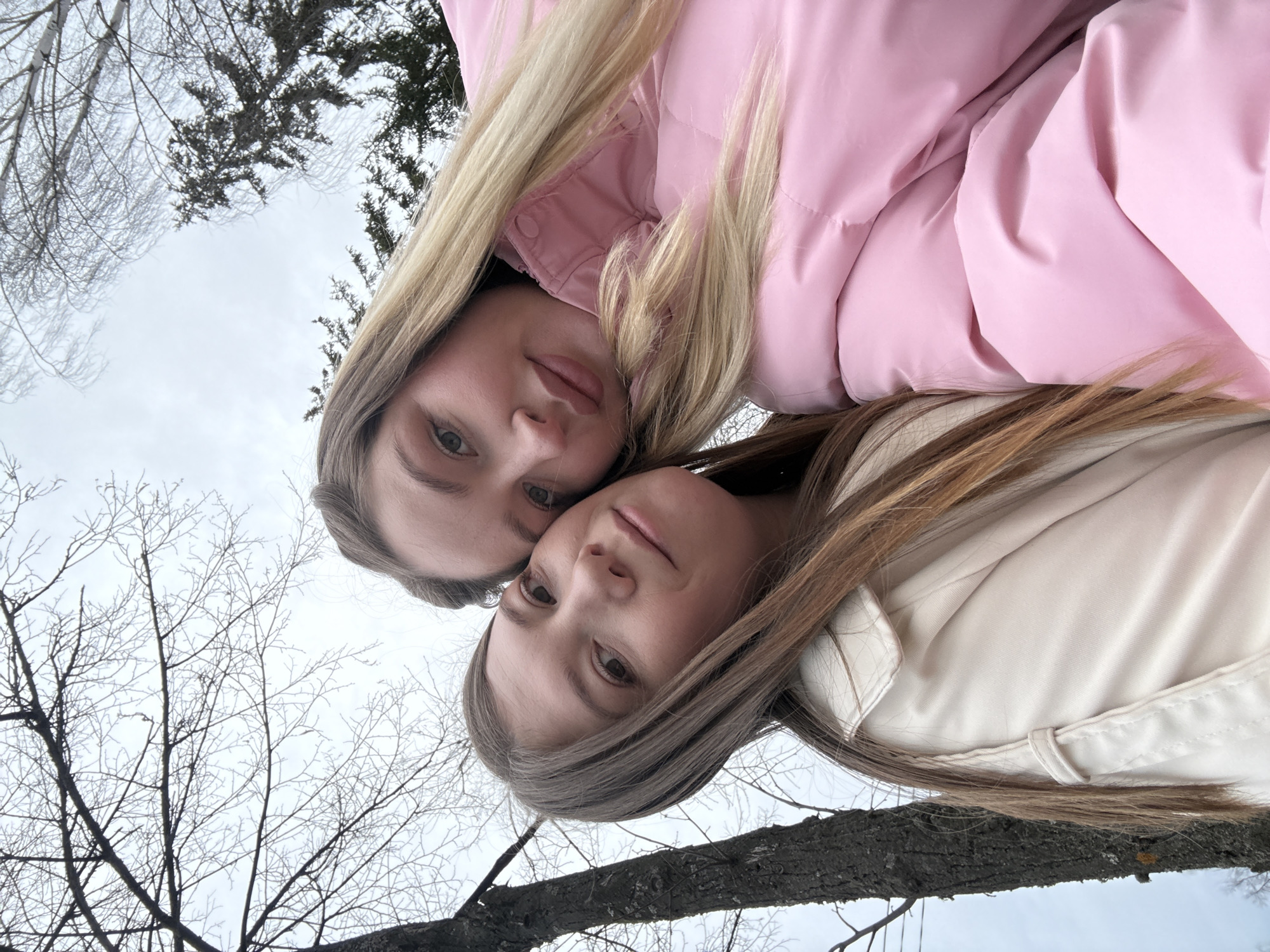 Two young women pose closely together outdoors. One woman with blonde hair wears a pink puffer jacket. The other woman has light brown hair and wears a light beige jacket. They appear to be in a park or wooded area in Chaikovsky, Russia, as bare trees are visible in the background. The sky is overcast.