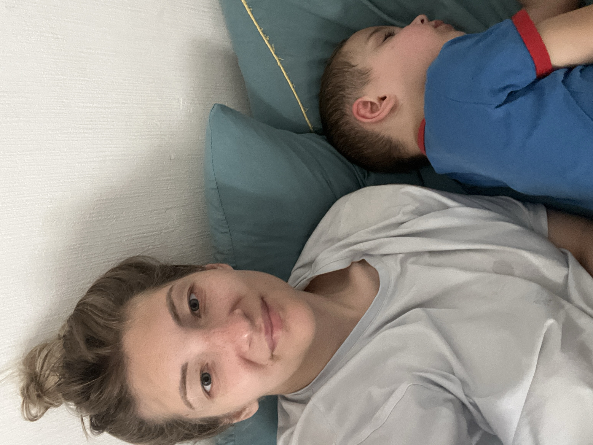 This is a selfie taken from a high angle, featuring two people lying down on a bed. On the right side of the image, a young boy is asleep, resting his head on a teal-colored pillow. He is wearing a blue t-shirt with red trim on the sleeves. His eyes are closed, and his face is relaxed in sleep. To the left, a woman is also lying down, looking towards the camera. She has her hair tied back in a bun and appears to have freckles on her face. She is wearing a light gray t-shirt. The background consists of a textured white wall and the teal pillow. The lighting suggests it might be daytime, and the overall mood is one of intimacy and relaxation, possibly during nap time or bedtime. The location is identified as Sochi, Russia. There is no visible text in the image.