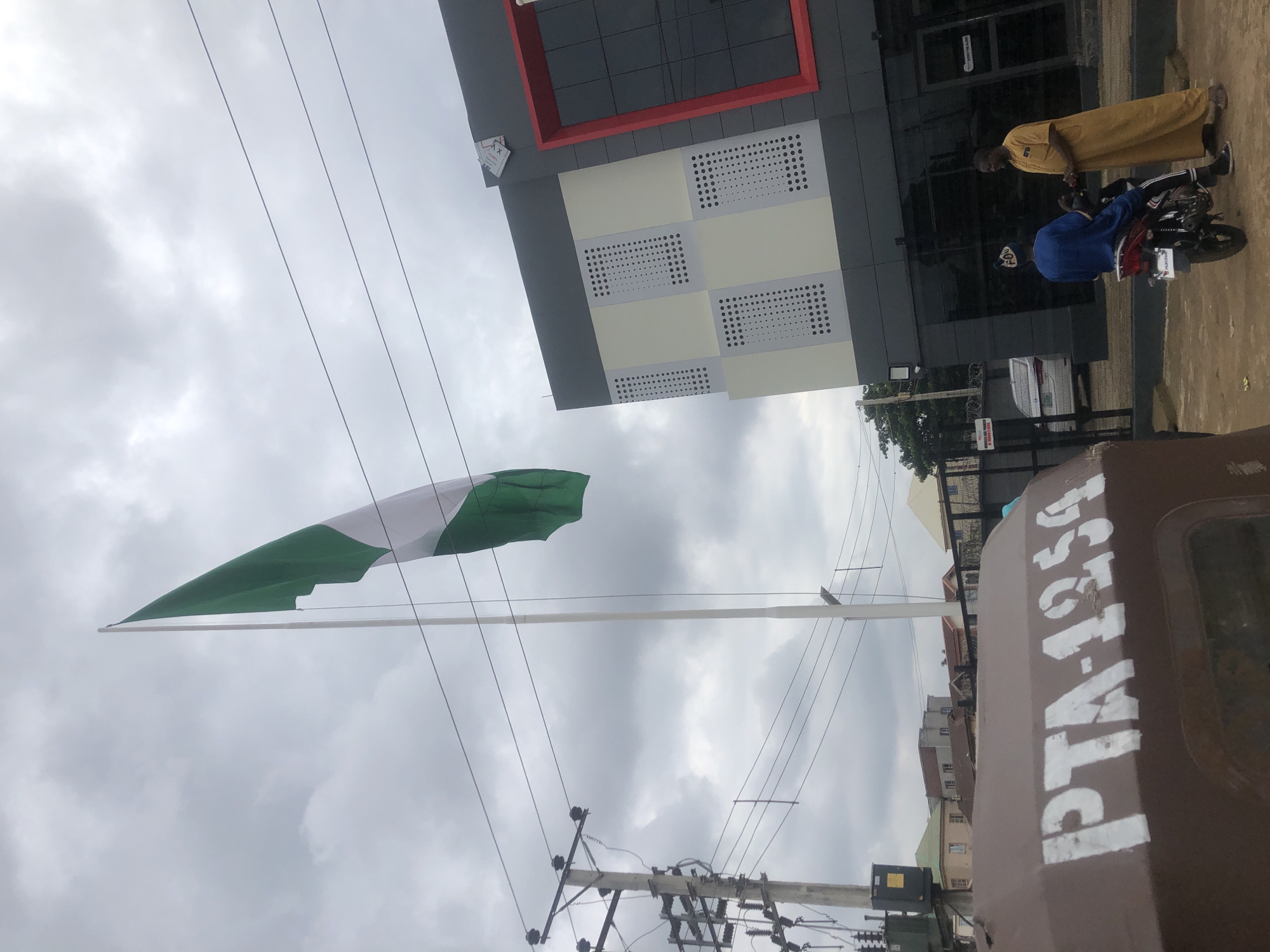 In Jos, Nigeria, an overcast sky looms over a street scene featuring a towering flag pole with the Nigerian flag, a two-tone green and white banner, flying prominently. To the right, a modern building with a gray facade, white paneling with perforated details, and a striking red frame around a dark glass window stands out. Two men are present; one, seated on a motorcycle, wears a blue outfit and a patterned cap, while the other, standing, is dressed in a flowing yellow robe. In the foreground, a brown vehicle with the stenciled white lettering "PTA-1254" is visible, its surface weathered. A utility pole with electrical equipment and wiring is on the left, and in the background, residential buildings with various roof colors are apparent. A fence, possibly topped with barbed wire, encloses some of the property.