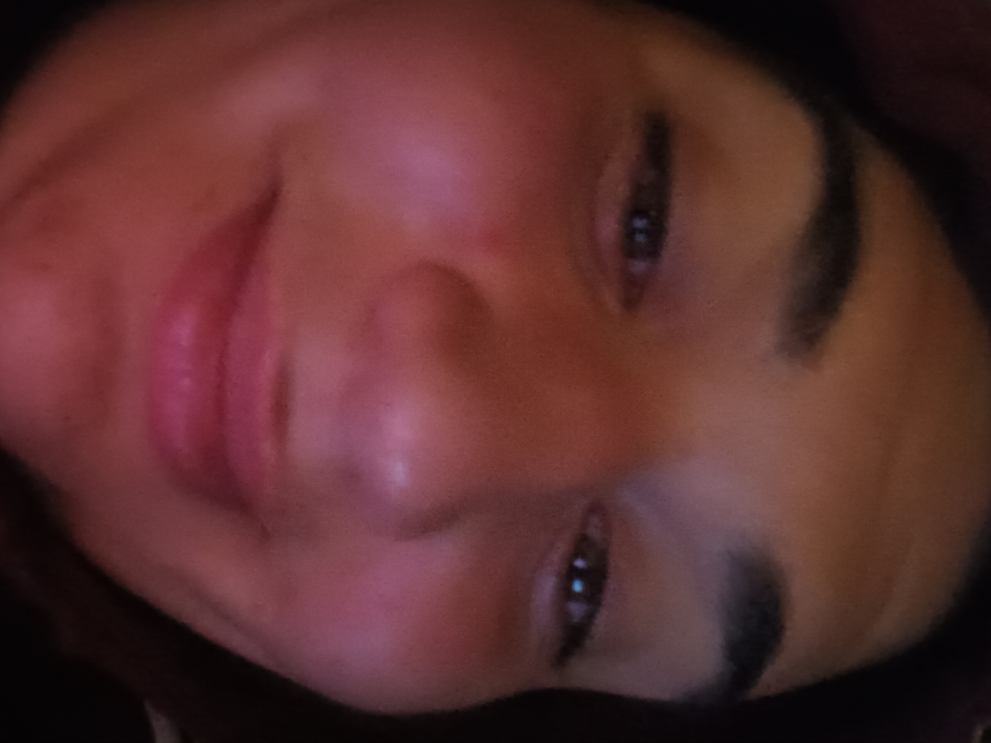 A close-up, slightly angled portrait of a woman's face. Her eyes are open, and she has dark eyebrows. Her lips are closed. The background is dark and indistinct. The location is unknown.