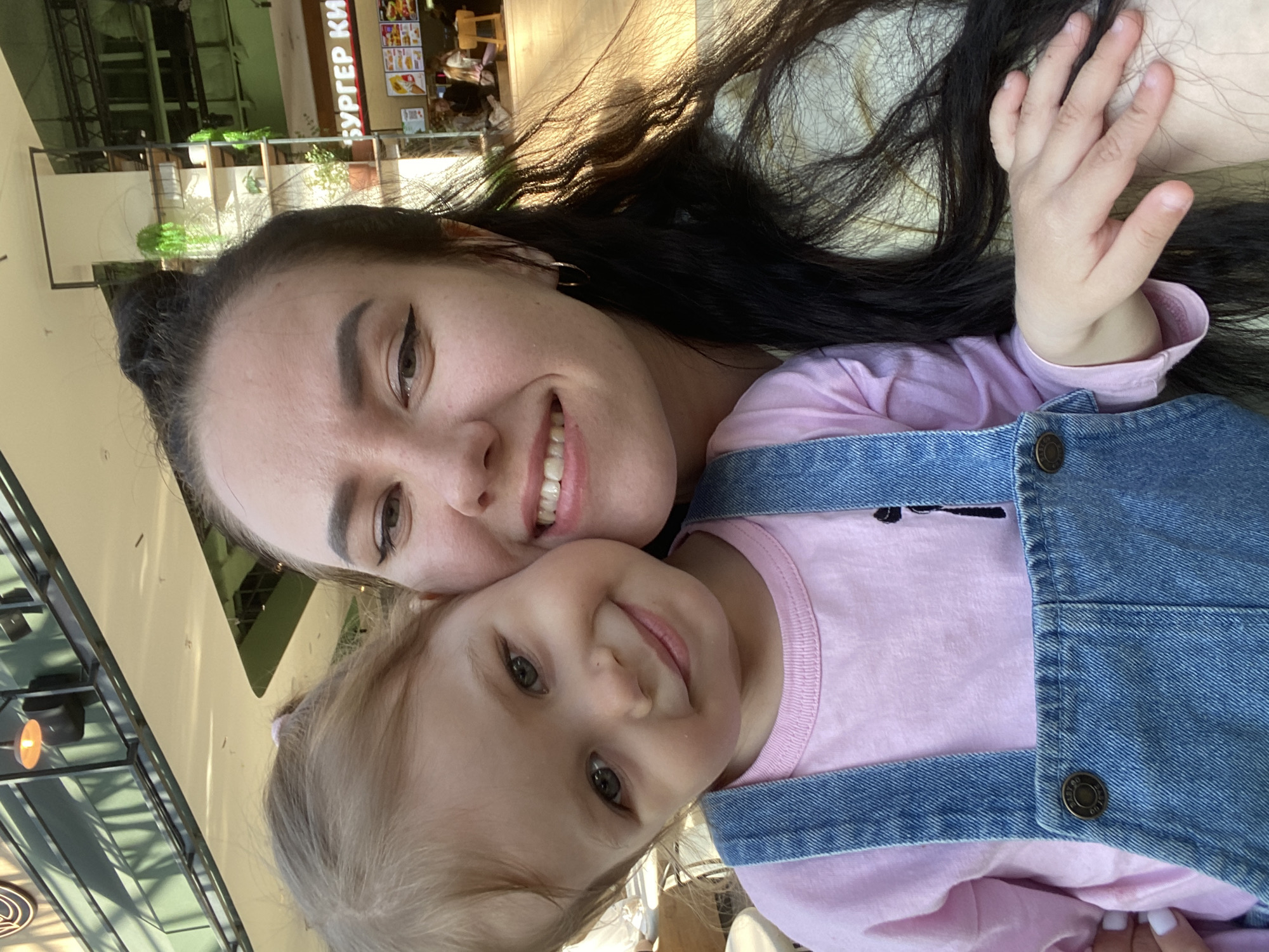 A woman and a child are posing for a selfie inside a building, likely a mall or shopping center, in Perm, Russia. The woman has long, dark hair and is smiling. The child, who appears to be a toddler, has light hair and is wearing a pink shirt under denim overalls. The background shows the interior of the building, with signs in Russian indicating a Burger King restaurant and other shops. The perspective suggests the photo was taken at an angle.
