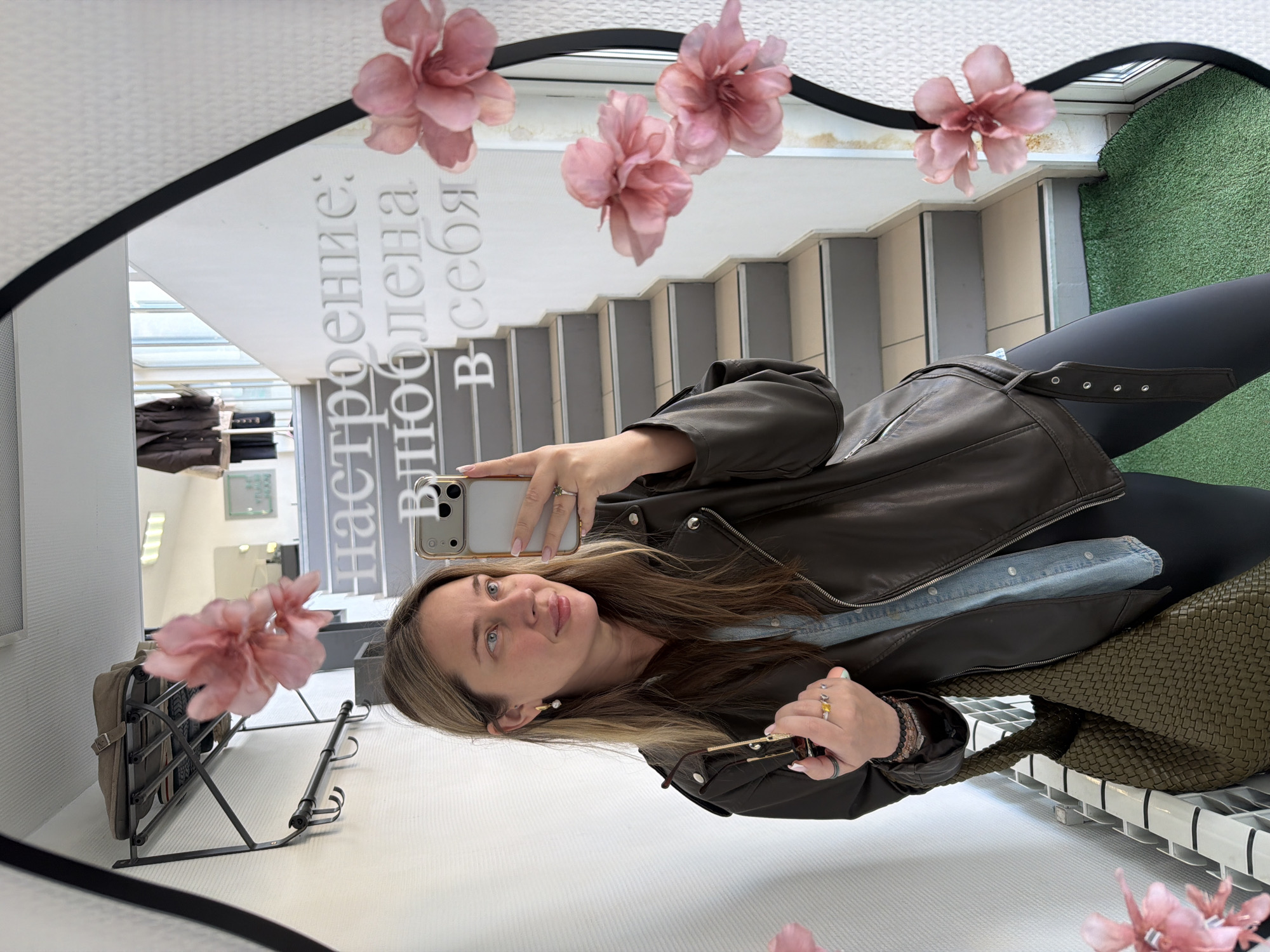 A woman is depicted taking a mirror selfie in an indoor setting, likely in Chaikovsky, Russia. She is shown from the waist up, holding a light-colored smartphone in her left hand to capture the image, and sunglasses in her right. Her light brown hair with highlights frames a face with light-colored eyes, and she looks directly at the camera with a gentle, slightly smiling expression. She is dressed in a dark brown leather jacket worn over a light blue denim shirt and black leggings. A dark green woven bag rests to her right. Her hands feature multiple rings, and she wears an earring in her visible ear.
The mirror she is using is adorned with several artificial pink flowers, giving it a decorative touch. Reflected in the mirror is a modern staircase with light-colored steps leading upwards. Other reflections include a clothing or shoe display rack and a patch of artificial green turf, possibly part of an interior design feature or an outdoor area visible through a window. Prominently visible on the mirror's surface is Cyrillic text, which partially reads: "настроение: влюблена в себя," translating to "mood: in love with myself." The setting appears to be a well-lit commercial or public indoor space during the daytime.
