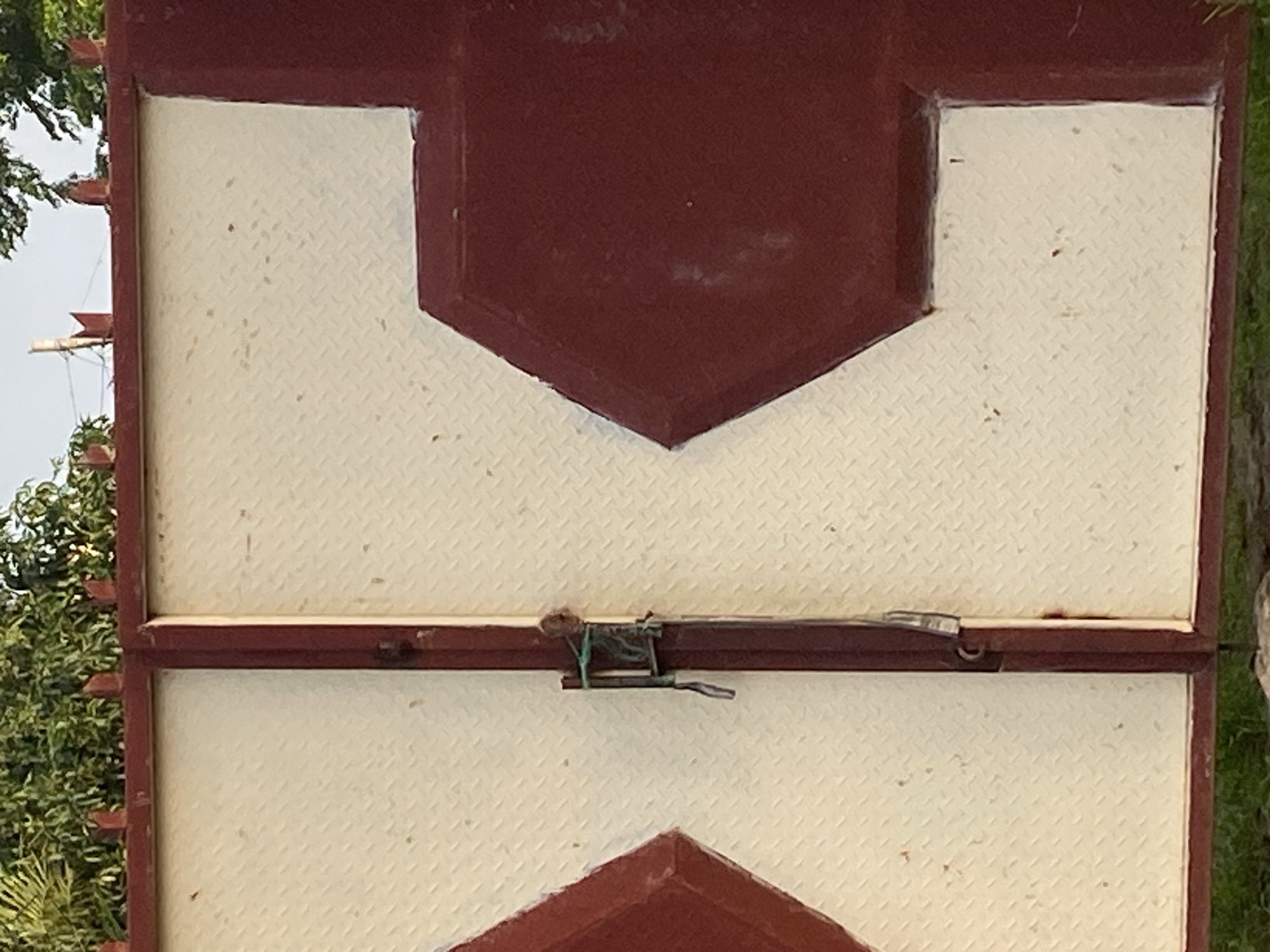 The image shows a close-up view of a closed gate in Jauro Yinu, Nigeria. The gate is made of metal and is painted a maroon color on the frame and a cream color on the panels. The cream panels have a diamond plate texture. There is a latch mechanism on the left gate, secured with what appears to be green twine. The top of the gate is adorned with pointed finials, and behind the gate, lush green trees are visible. The ground in front of the gate is covered with grass.