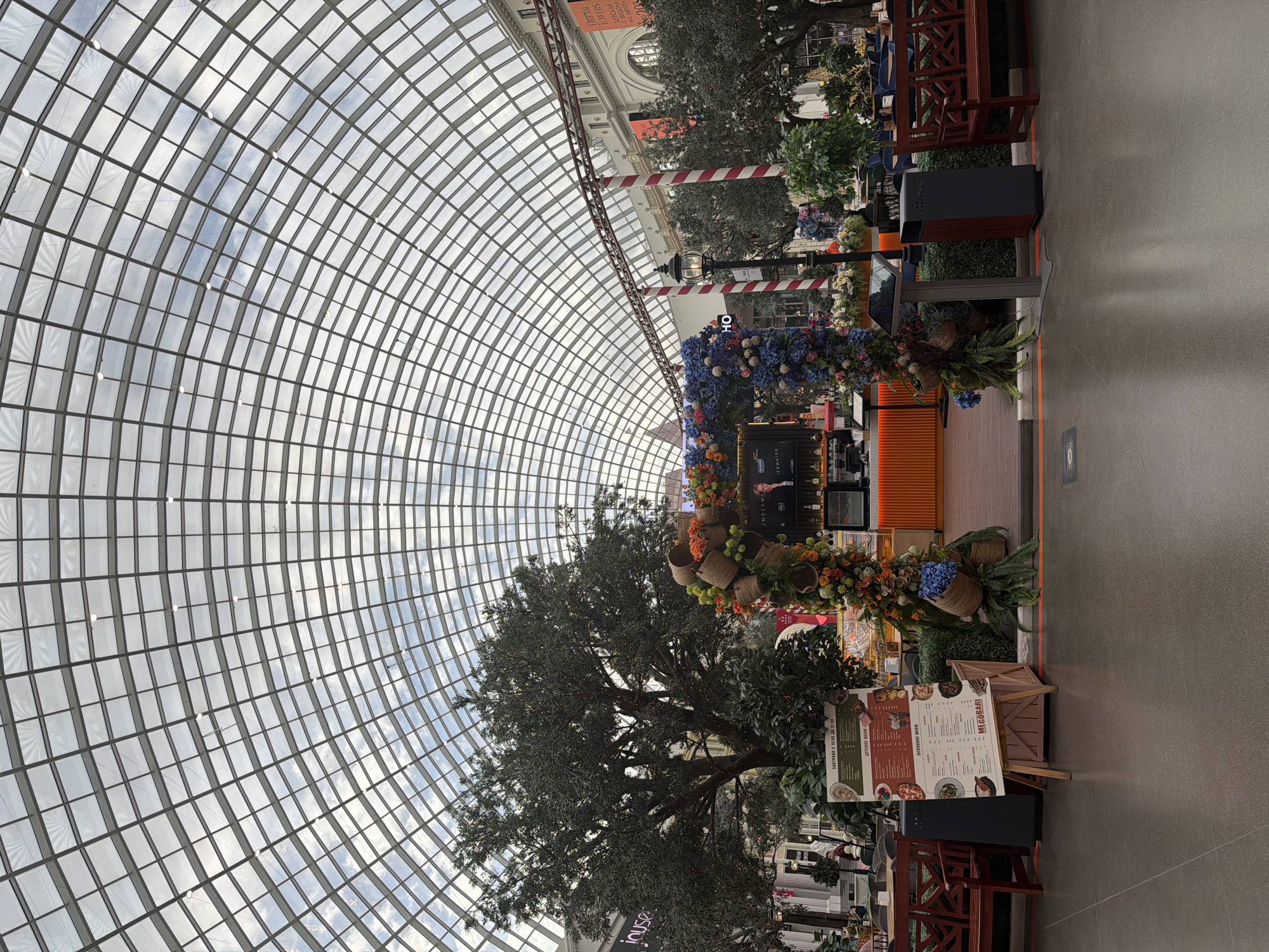 The image shows the interior of a large, enclosed space with a curved glass ceiling. In the foreground, there is a decorative archway adorned with flowers and baskets. To the left of the archway, a wooden bench and a sign stand on the tiled floor. In the center of the image, a large artificial tree is visible. Beyond the archway, an orange counter with a display of drinks is present. The background reveals more seating areas, greenery, and architectural details of the building. The scene appears to be a public space, possibly a shopping mall or an atrium.