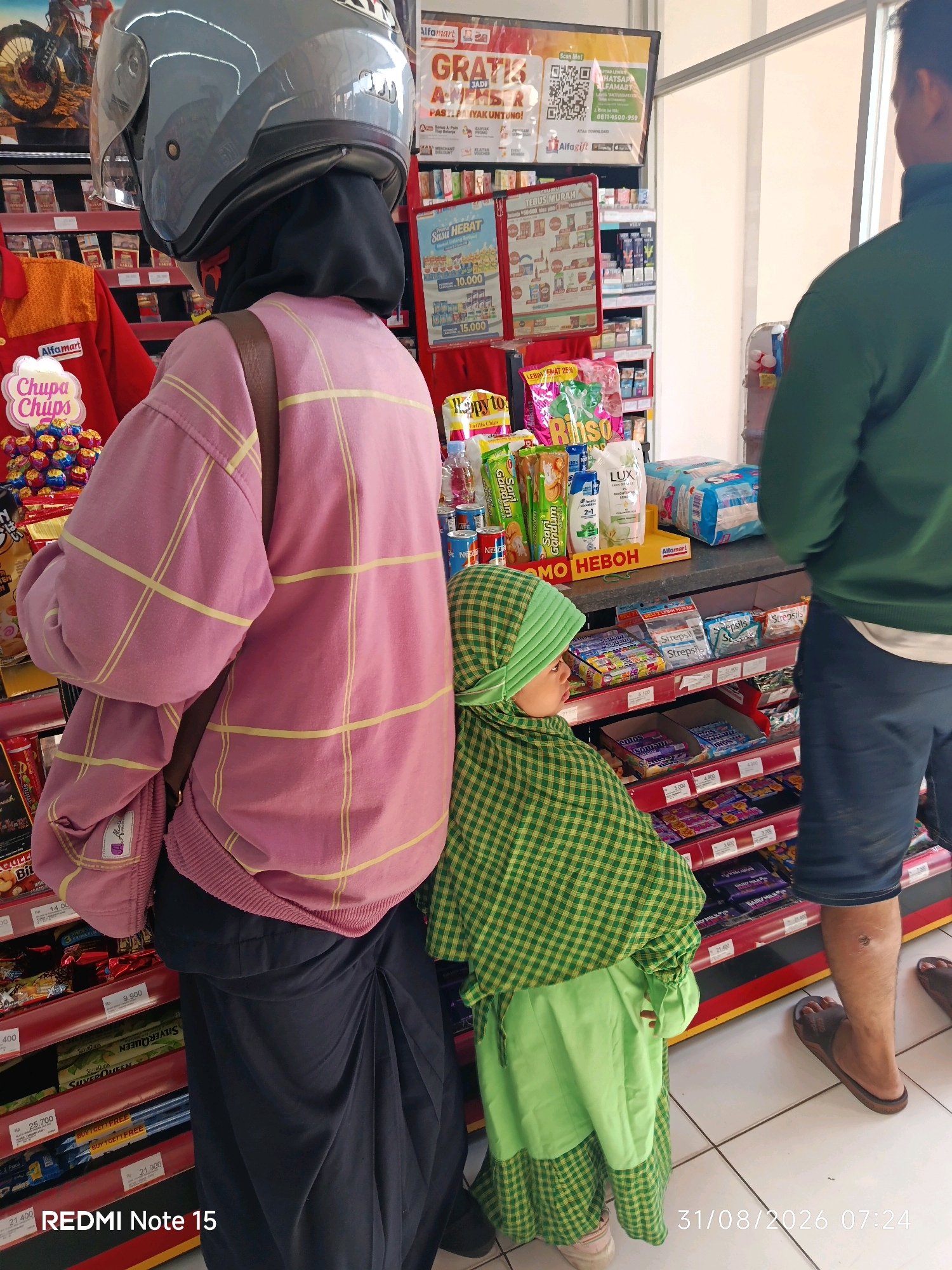 A full-shot, eye-level interior photograph shows a mother and child in an Alfamart convenience store in Keboansikep, Indonesia. The mother, wearing a pink plaid oversized sweater, black hijab, and black skirt, stands facing away from the camera. She wears a grey helmet. A younger child, wearing a green plaid hijab and dress, stands to the mother's right, facing the store's shelves. A man in a green sweater and blue jeans stands to the right of the child, also facing the shelves. The shelves are stocked with various food and household items, including candy, diapers, and toiletries. The store's interior is brightly lit with fluorescent lights. The floor is tiled in a white checkerboard pattern. The date and time on the bottom right of the image read "31/08/2026 07:24". The bottom left corner has text "REDMI Note 15".