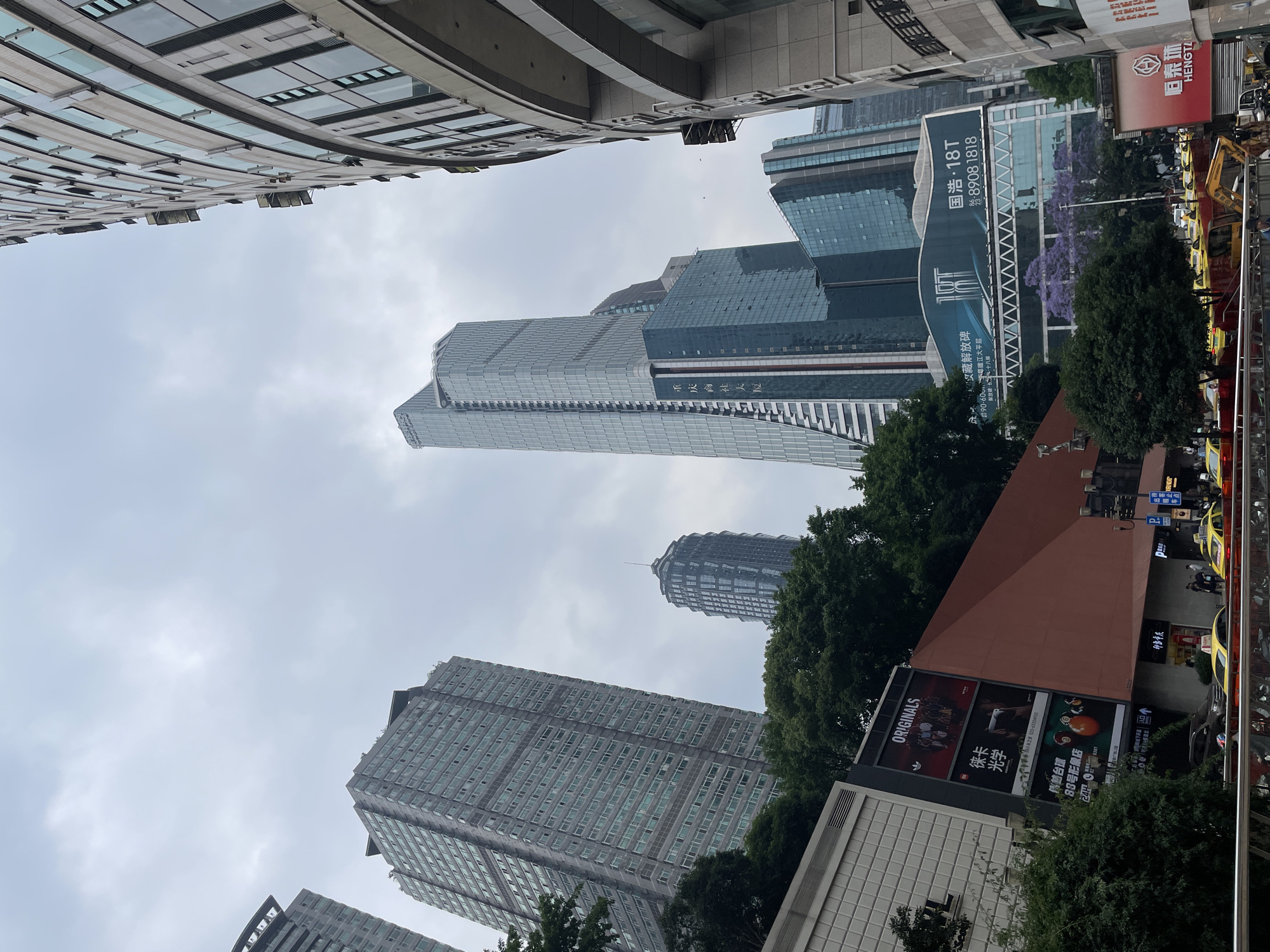 The image depicts a street view in the Yuzhong District of Chongqing, China, characterized by a high density of modern high-rise buildings. In the foreground, there is a commercial building advertising "Originals" and "Leica Optics" which fronts a street with several parked yellow taxis. Looming above and behind this building, the perspective shifts upwards to reveal a cluster of sleek skyscrapers, partially obscured by green trees. The sky is overcast, with a pale, uniform cloud cover. The buildings feature modern glass facades, and one structure shows an advertisement banner for "Guohao 18T". The perspective is tilted upwards, emphasizing the scale of the architecture within a dense urban environment.