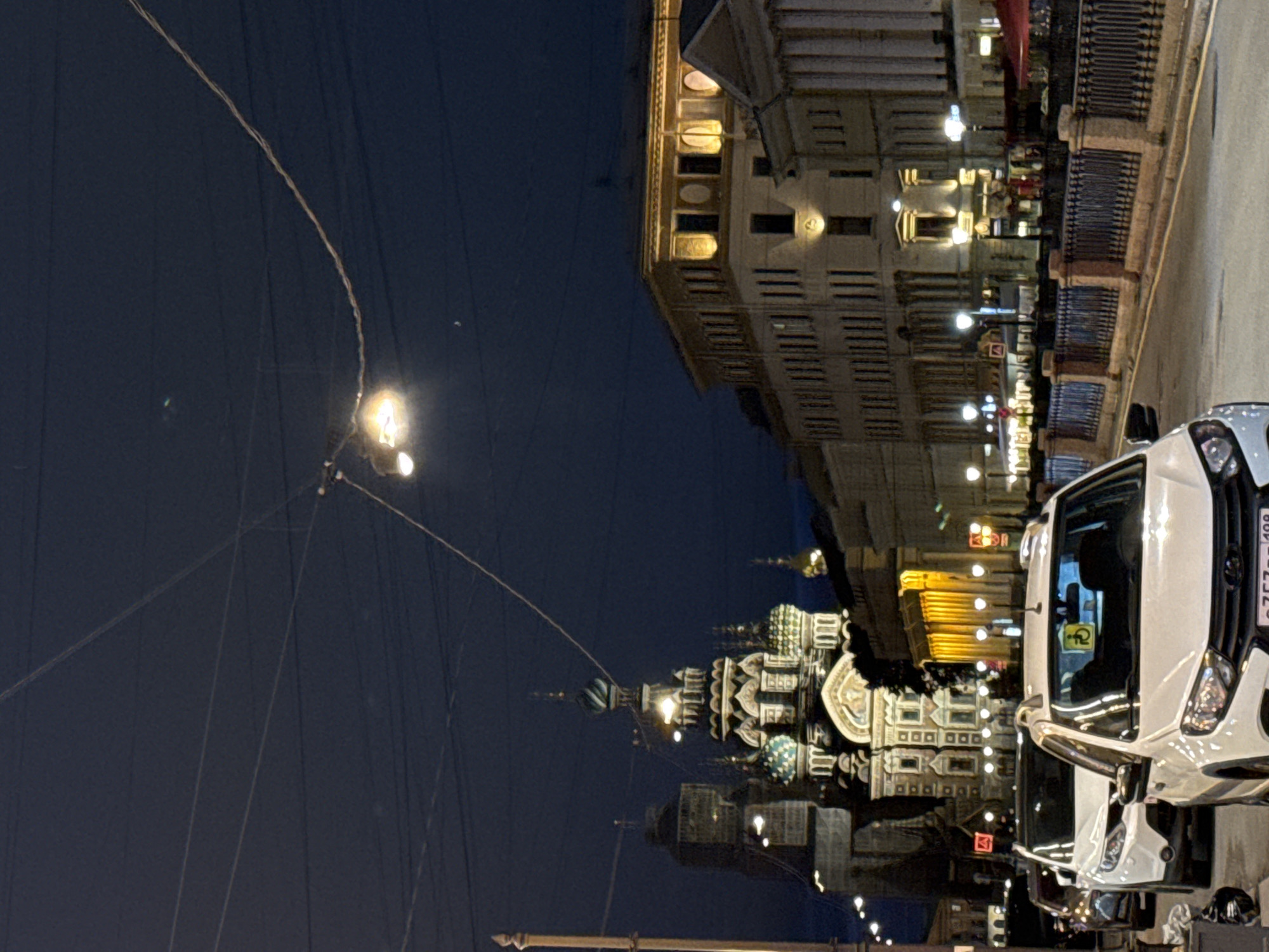 In Saint Petersburg, Russia, at night, the Church of the Savior on Spilled Blood is prominently featured. It is illuminated and adorned with numerous domes. To the right of the church, a row of buildings with illuminated windows and ornate architecture lines a street. A metal fence separates the sidewalk from a body of water, likely a canal, which is not visible. Several cars are parked on the street, including a white car in the foreground with a disabled parking permit sticker on its windshield. Overhead, power lines and streetlights crisscross the dark sky. The scene depicts a city street at night, highlighting historical architecture and modern vehicles.