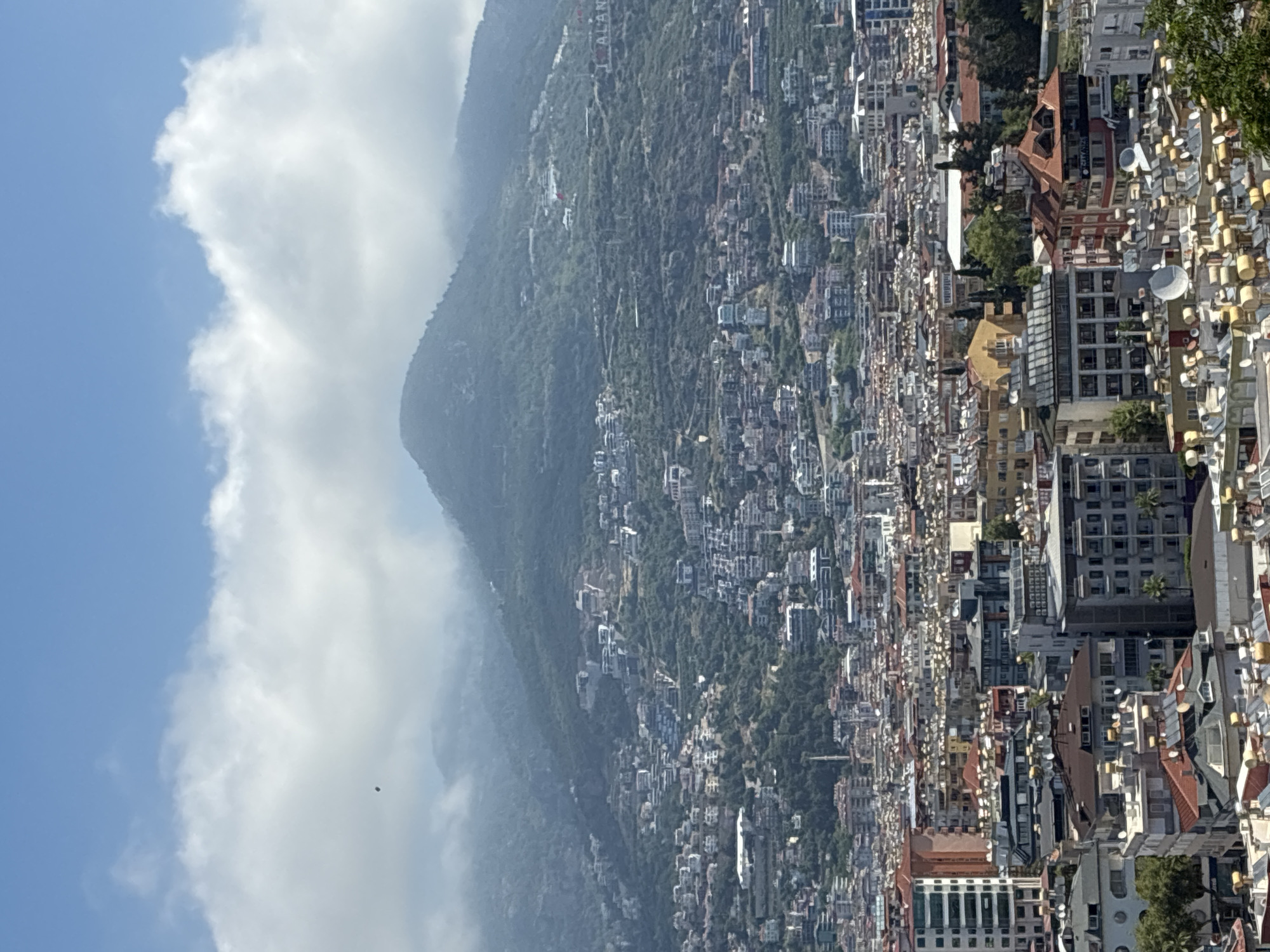 This image captures an expansive, elevated view of Alanya, Turkey, showcasing a dense urban landscape nestled against a large, verdant mountain. The scene is presented from a high vantage point, looking across the city towards the natural backdrop.
The city occupies the middle and foreground, characterized by a multitude of buildings packed closely together. These structures display diverse architectural styles, with many featuring light-colored facades and a mix of flat or reddish-brown tiled roofs. Numerous satellite dishes are visible on rooftops, indicative of a modern, developed area. The urban sprawl extends from a flatter coastal area and gradually ascends the lower slopes of the mountain.
In the background, a massive mountain rises majestically, its slopes densely covered with lush green vegetation, suggesting a heavily forested or scrubland environment. White, fluffy clouds partially shroud the mountain's peak and trail along its upper ridges, creating a dynamic atmospheric effect against the clear blue sky visible above. The bright lighting confirms it is daytime.
A prominent identifying feature is visible on the mountain's slope: large white letters forming the phrase "I ❤️ ALANYA," with a red heart symbol or flag nearby. This serves as a significant landmark and firmly places the scene in Alanya. On a building within the city, the text "CITYMATE" is partially discernible on its side.
The image portrays a serene yet vibrant urban-natural interface, with no discernible human activity taking place, focusing instead on the impressive panorama of the city and its mountainous surroundings under a partly cloudy, bright sky.