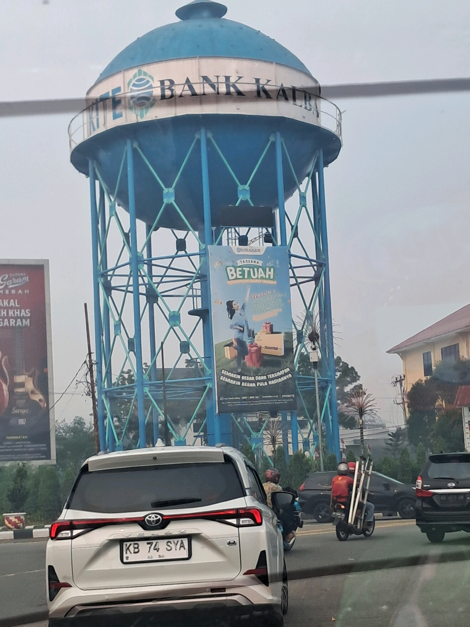 The image depicts a street scene in Pontianak, Indonesia, dominated by a large, blue water tower with the words "BANK KALTIM" visible near the top. The tower has a metallic, lattice-like structure supporting a spherical reservoir. A large banner hangs from the tower, featuring text and images related to "TASERNA BETUAH," possibly an advertisement. On the left, a billboard advertising "GARAM MERAH" (red salt) with images of guitars is partially visible. In the foreground, the rear of a white Toyota Veloz is captured, displaying the license plate "KB 74 SYA." A motorcycle carrying a long ladder is visible in the middle ground, along with other vehicles and pedestrians. The environment appears to be an urban setting with buildings and trees in the background. The sky is overcast, suggesting it might be a cloudy day.