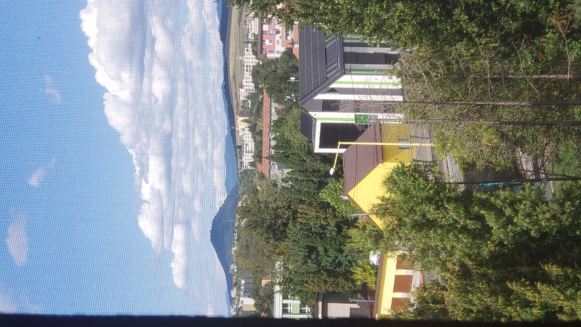 This is a view from a window overlooking a city. In the distance, a large, verdant mountain dominates the skyline under a blue sky with scattered white clouds. Below the mountain, a cityscape is visible with various buildings, including residential blocks and some smaller structures. In the foreground, there are trees and several modern buildings. One building in the foreground is yellow with a brown roof, and next to it is a contemporary building with a sloped roof covered in solar panels, featuring vertical stripes of white, green, and black. The view is partially obstructed by a window screen. This scene is likely from Prešov, Slovakia, as indicated by the characteristic landscape and architecture.