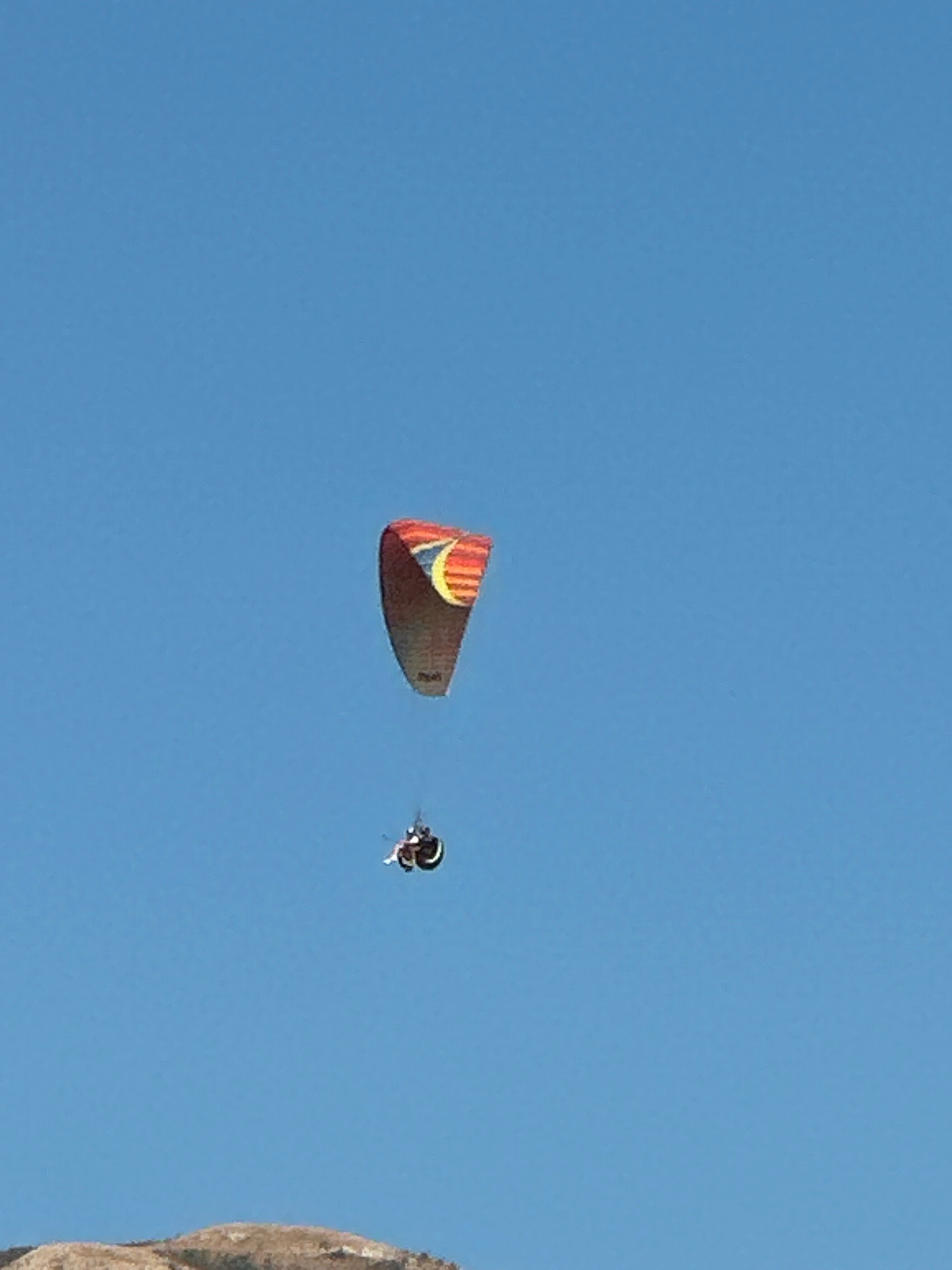 A paraglider is seen soaring through a clear blue sky above Boreti, Montenegro. The paraglider's wing is a vibrant mix of red, orange, and yellow stripes, with a yellow crescent moon design. A pilot is suspended beneath the wing in a harness, appearing to be in mid-flight. The paraglider is positioned in the upper right quadrant of the image. In the lower portion of the image, a portion of a sandy, rocky hill is visible, likely part of the landscape near Boreti. The overall scene depicts a recreational activity taking place in an outdoor, natural environment.
