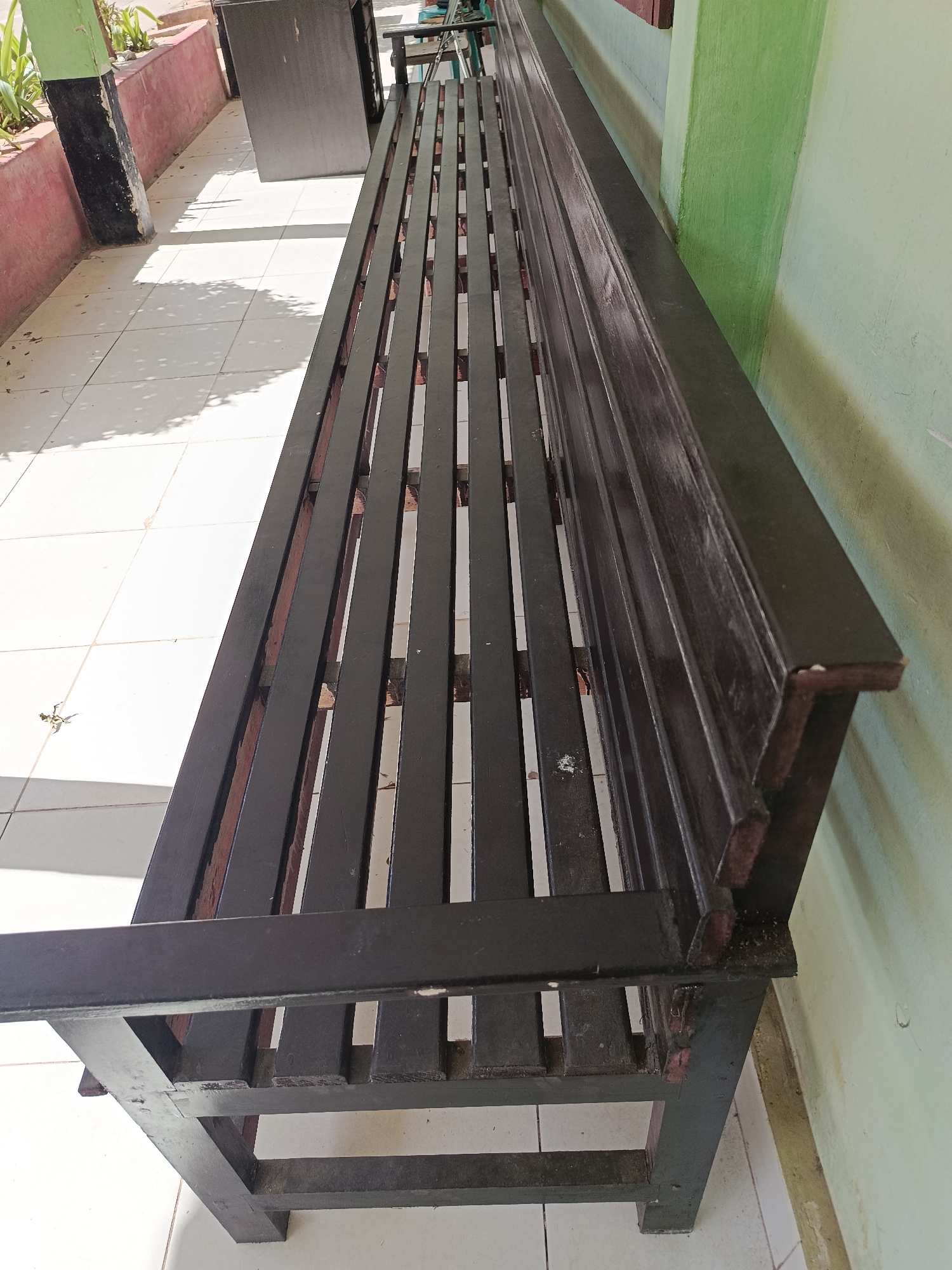A long, dark brown wooden bench with a slatted seat and backrest sits on a white tiled floor. The bench is positioned alongside a pale green wall. To the left of the bench, a portion of a red and black concrete pillar is visible, with some green plants growing behind it. Sunlight casts shadows on the tiled floor, suggesting an outdoor or semi-outdoor location, possibly a public space or entryway in Mansalean, Indonesia. There are no people or animals present in the image. The bench appears to be aged, with some wear and tear visible on its surfaces and edges.