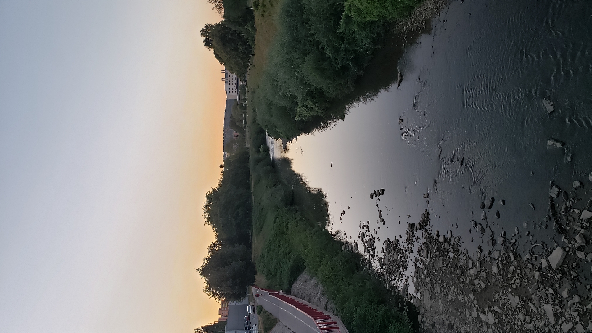 The image depicts a tranquil scene along the Torysa River in Prešov, Slovakia, likely during the late afternoon or early evening due to the soft, warm light illuminating the sky. The river, characterized by its calm flow and rocky bed, meanders through a verdant landscape. On the left bank, a paved walkway with a red railing follows the river's course, with a single cyclist visible, indicating a recreational area. Adjacent to the walkway, a steep embankment is covered in dense green vegetation. Across the river, the banks are also lined with thick bushes and trees, some of which are quite tall. In the distance, beyond the trees, buildings and structures are visible, suggesting the presence of a town or city. The sky transitions from a pale blue at the top to a soft orange hue near the horizon, typical of a sunset or sunrise. The overall impression is one of natural beauty integrated with urban development.