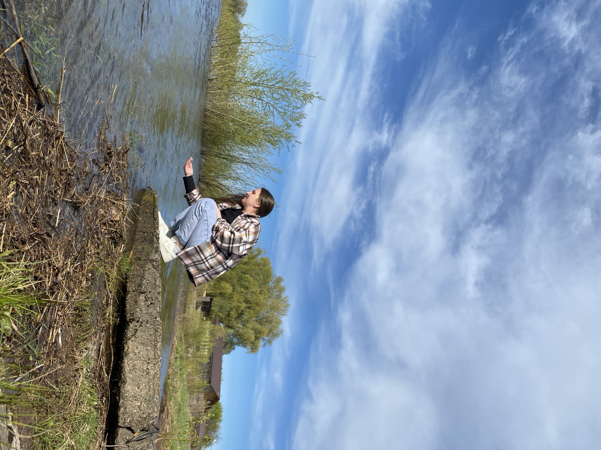 A young woman is crouching by the edge of a body of water, possibly the Kama River, in Chaikovsky, Russia, on a clear, sunny day with cirrus clouds in the sky. She wears jeans, a black top, and a plaid flannel shirt. Her hair is long and dark. Her right hand is extended, lightly touching the water. The edge where she sits is composed of grass, dead vegetation, and a low concrete or stone border. Behind her are trees and a building. The water is relatively calm.