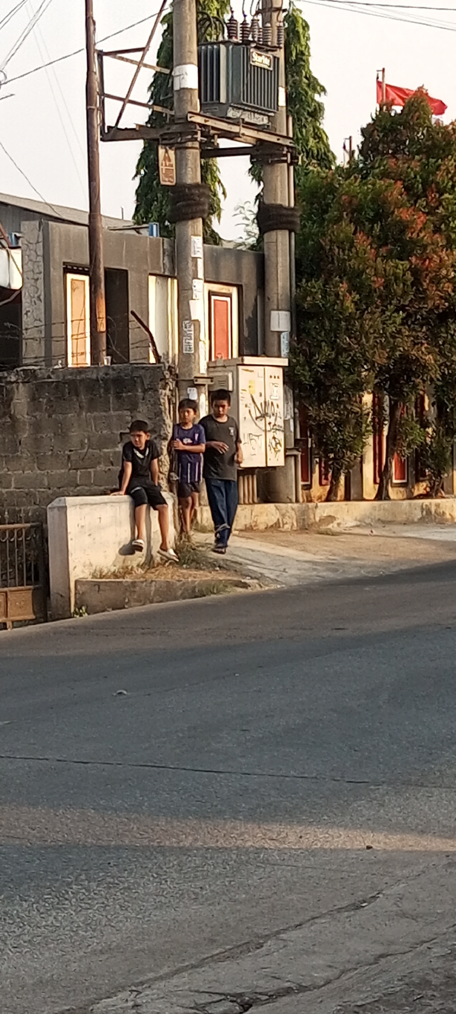This is a medium shot image captured outdoors in Setu, Indonesia, focusing on three young males standing and sitting near a utility pole and a low wall. The overall lighting suggests it is daytime, with shadows indicating the sun is at an angle.
In the foreground, a paved road with visible cracks and markings is present. To the left, a low, concrete wall or barrier runs horizontally across the frame. A young male, wearing a black t-shirt and black shorts, is seated on this wall, facing left. He has short dark hair and is wearing white sandals. To his right, a second young male stands, looking forward. He is wearing a short-sleeved shirt with purple and white stripes and dark shorts, with short dark hair. Further to the right, a third young male stands, also looking forward and slightly to the right. He is dressed in a dark, short-sleeved t-shirt and dark pants, with short dark hair. His body is angled slightly towards the viewer.
Behind the individuals and the wall is a multi-story building with a grey, textured exterior. Several doors with reddish-orange frames are visible on this building. To the right of the building, a tall utility pole with various components, including a transformer box and insulators, stands prominently. The pole has thick, dark bands wrapped around its base. Adjacent to the utility pole, a cluster of trees with dense green foliage and some reddish-orange leaves is visible, with a red flag flying from a pole above the trees on the right side of the frame.
The ground in front of the building and around the utility pole appears to be a mix of concrete and dirt with sparse vegetation. The scene depicts a casual outdoor environment in an urban or semi-urban setting.