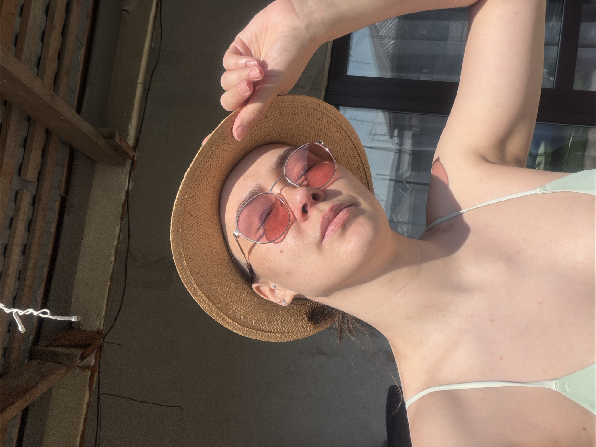 A woman with pale skin and freckles poses for a selfie. She wears a straw hat and pink tinted sunglasses. Her left hand rests on the brim of the hat. She also wears a light green top. Behind her, a window is visible, as well as wooden beams and a concrete wall, indicating an outdoor location possibly in Chaikovsky, Russia.