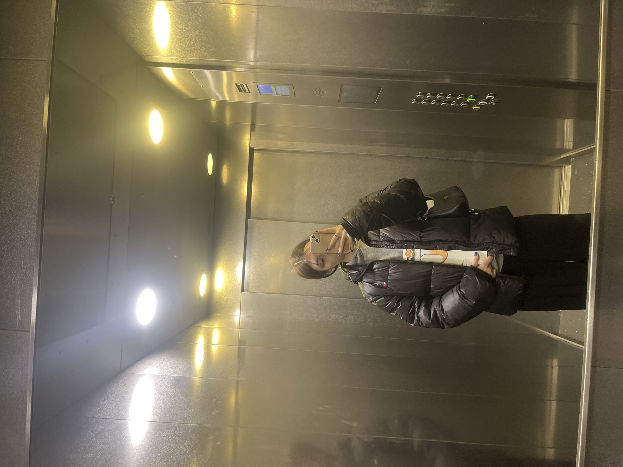 A person stands in a mirrored elevator and takes a selfie with a phone. The person is wearing a black puffer jacket, gray sweater, and black pants. The elevator has metal walls and a row of lights. There are elevator buttons above the person. This is a typical elevator found worldwide, and cannot be identified with Saint Petersburg, Russia.