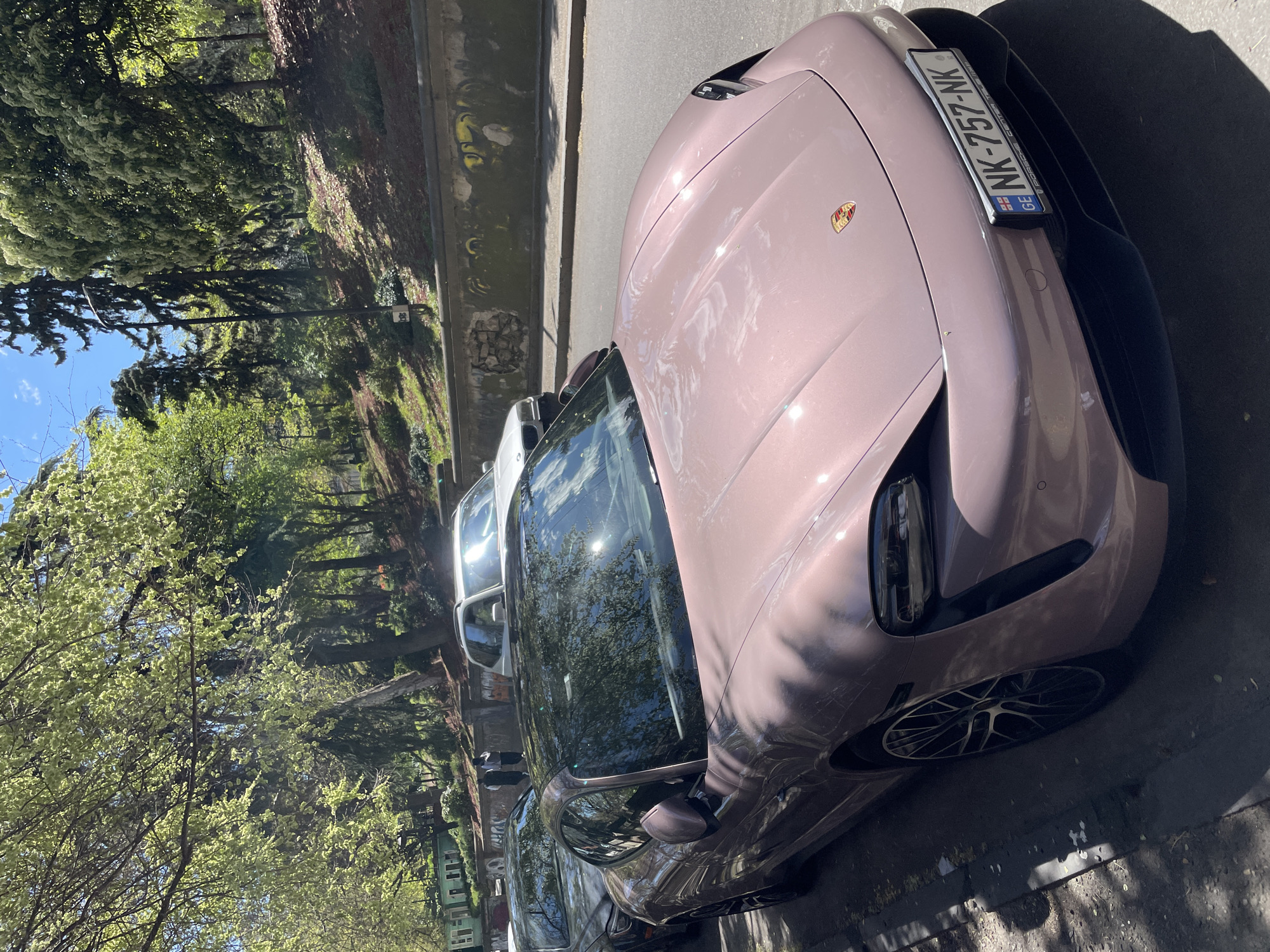 A parked Porsche, light purple or mauve in color, is prominently featured in the image. Its license plate reads "NK-757-NK", with "GE" indicating Georgia. The car is parked on a city street alongside other vehicles. The surrounding environment appears to be urban, with trees lining the street. Based on the license plate, this scene is likely located in the city of Tbilisi, Georgia.