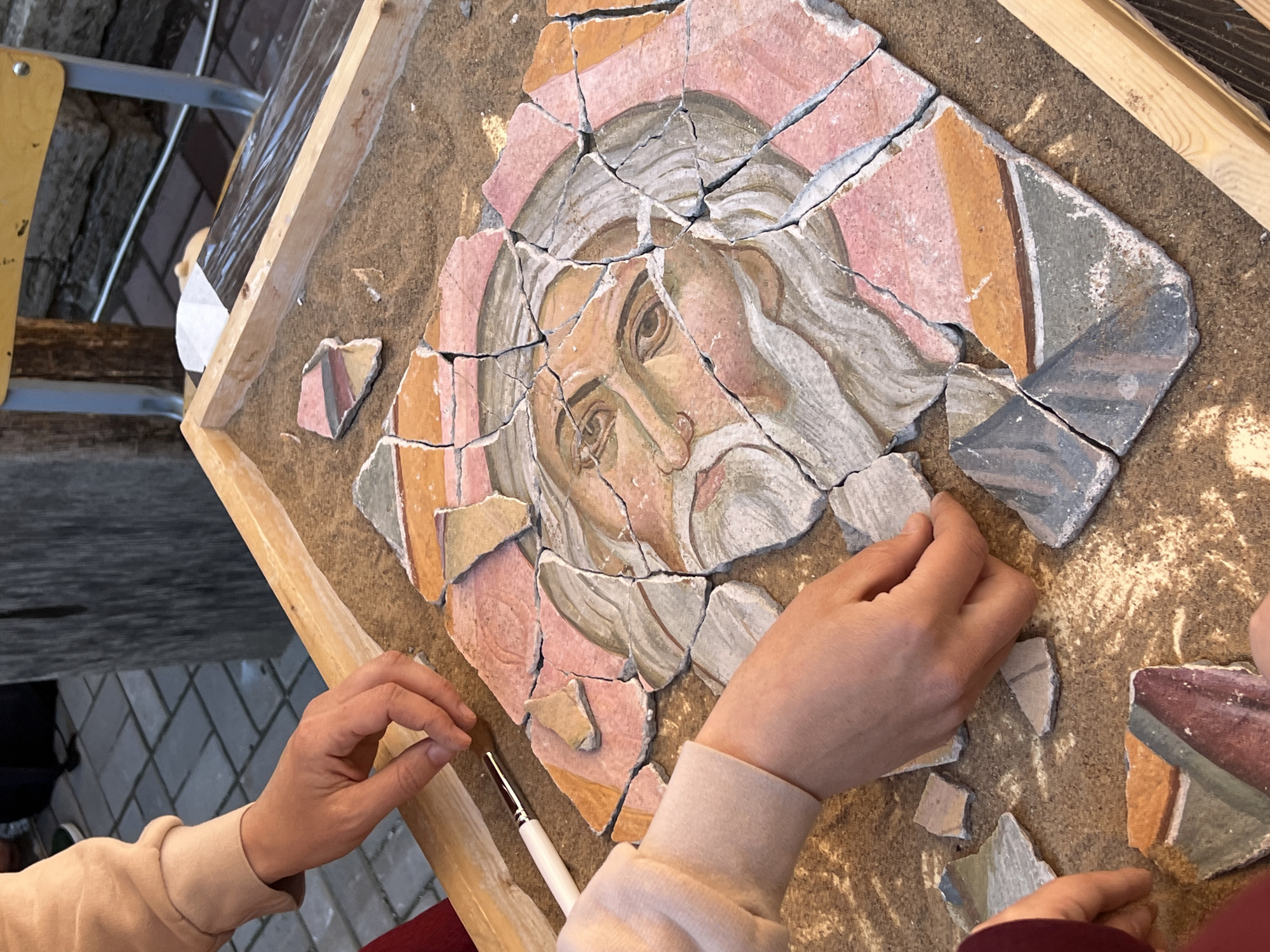 The image shows a fragmented Byzantine-style mosaic featuring the face of a bearded figure with white hair and beard. The mosaic is being reconstructed by human hands. The fragments are laid out on a flat, wooden-framed surface. A small brush is visible near the hands. The mosaic's color palette includes pink, tan, grey, brown, and white. The image has a forensic appearance with a focus on the mosaic and the reconstruction process. Based on the artistic style and the presence of mosaic art, the location could potentially be within a museum or conservation workshop in Saint Petersburg, Russia, but this cannot be determined with certainty from the image alone.