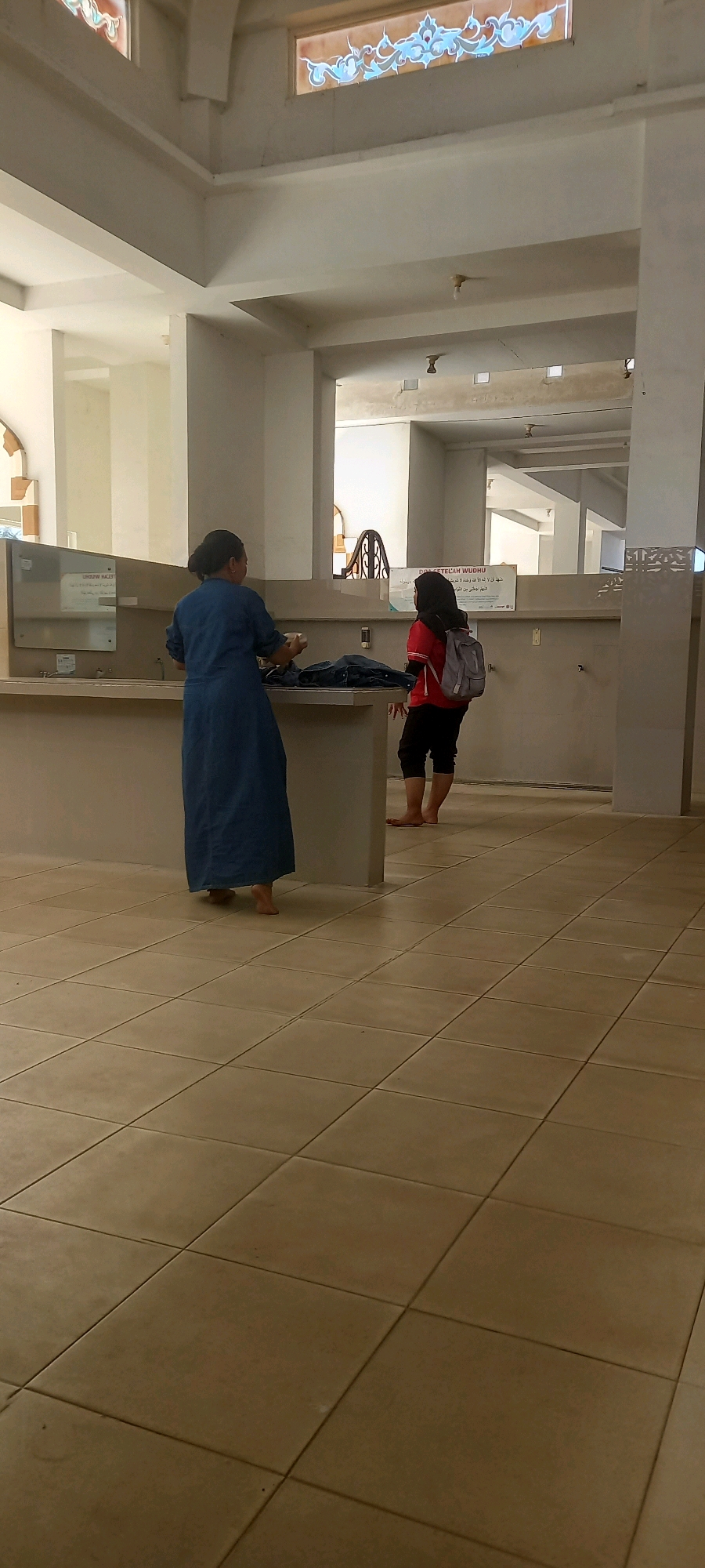 Two individuals are present in what appears to be a public restroom or ablution area in Kaliwates, Indonesia. The floor is tiled with large, square, beige tiles. In the foreground, a person wearing a long, dark blue dress is standing with their back to the camera, facing a counter. They are holding what appears to be a small white object in their hands. In the middle ground, another person wearing a red shirt and black pants is standing with their back to the camera and their right side facing forward. They have a grey backpack slung over their left shoulder and are barefoot. A sign with Arabic script is visible on the wall behind them. The environment is enclosed, with walls and a ceiling visible. The lighting appears to be natural, possibly from windows or open areas.