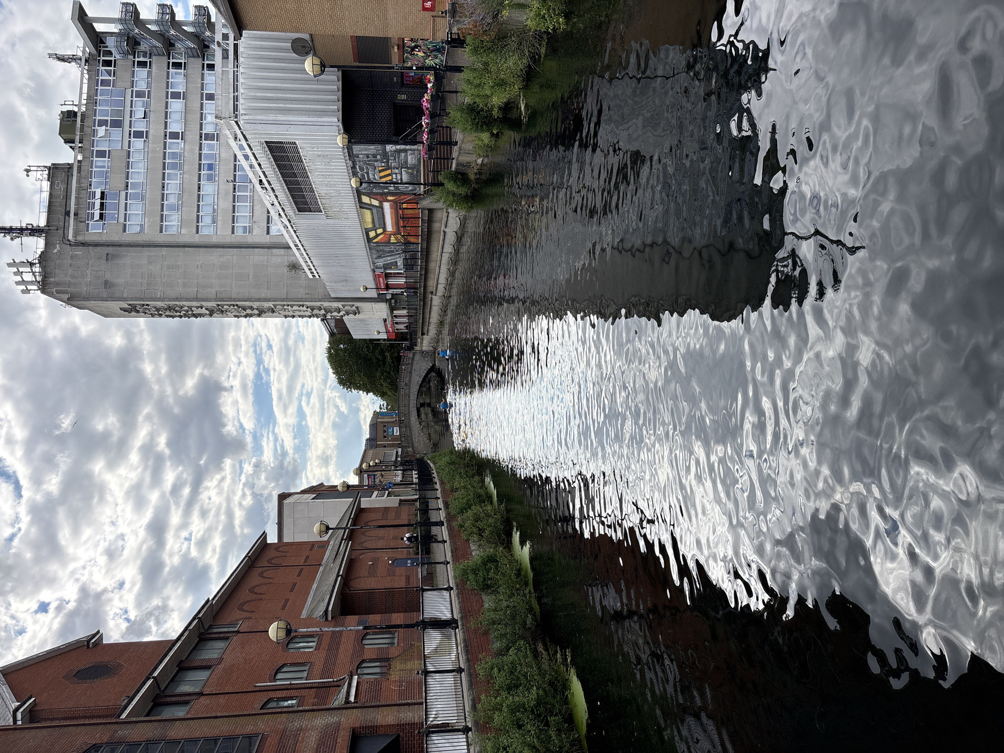 In the city of Unknown, Unknown, a canal runs through an urban landscape. The canal's surface is rippled, reflecting the overcast sky and the surrounding buildings. On the left bank, a red brick building with arched windows and a rounded tower is visible. A black metal railing runs along the embankment, bordered by green bushes. A single person is seen walking on the pavement. On the right bank, a tall, grey, multi-story building with many windows dominates the scene. Below it is a modern structure with a white ribbed facade. The canal is spanned by a stone bridge in the distance, where two figures are seen in small boats. The overall impression is one of a calm, urban waterway with a mix of old and new architecture.