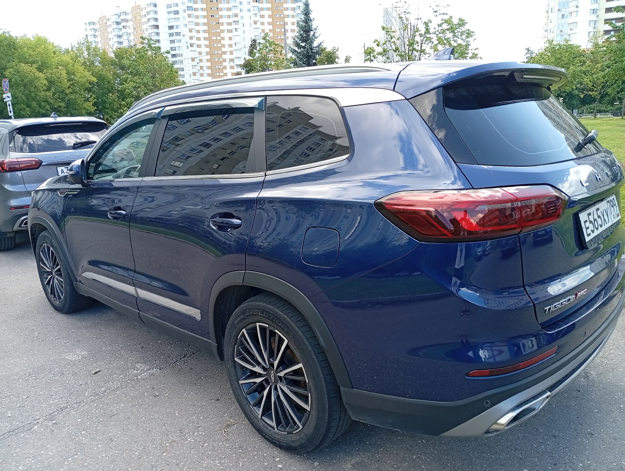 In the city of Unknown, Unknown, a dark blue SUV is parked on an asphalt surface next to a grassy area. The SUV is seen from its rear passenger side, with the front of the vehicle out of frame to the left. A silver SUV is parked behind it. In the background, tall, multi-story buildings are visible, along with trees and a clear sky. The license plate on the dark blue SUV is partially visible and reads "E565XY 799 RUS". The car has a sleek design with tinted windows and a rear spoiler. The overall scene suggests a residential or urban setting during daylight hours.