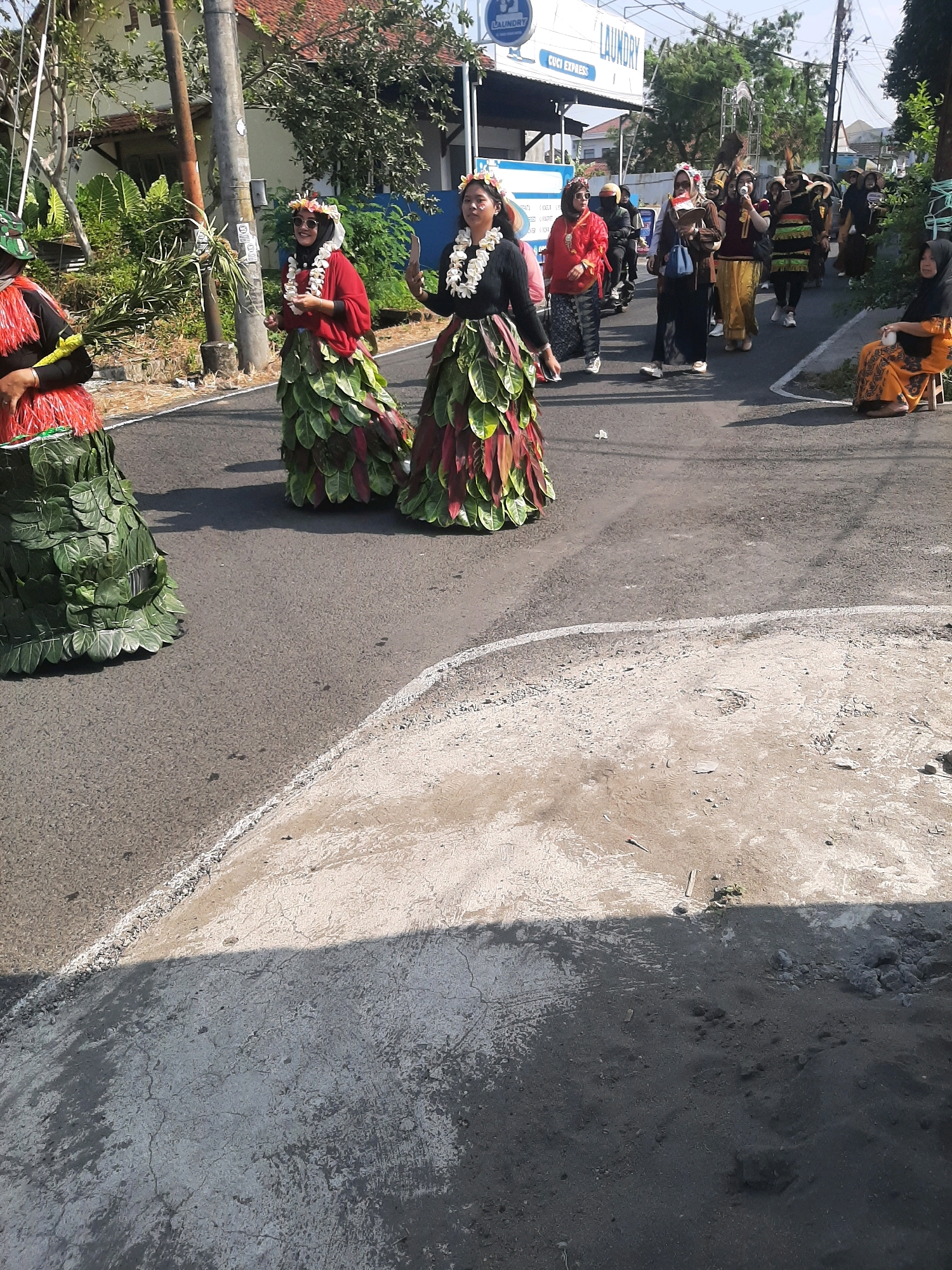 In Sumampir, Indonesia, on a sunny day, several women in elaborate costumes walk along a paved road. The women in the foreground are wearing dresses made of green and red leaves. The woman on the far left has a dress composed entirely of large green leaves and is wearing a green and brown camouflage headscarf. The woman next to her wears a red top and a skirt made of layered green and red leaves, adorned with a flower crown. The third woman from the left has a black top, a leafy skirt, and a flower crown. Behind them, a group of other women are walking, one in a red top and a patterned skirt, another in a brown top and gold pants. A woman in an orange and black patterned outfit is seated on a stool on the right side of the road. A laundry establishment with a "LAUNDRY" sign is visible in the background, suggesting an urban or commercial area. The road has a distinct white line marking its edge. The ground in the foreground is a mix of rough, unpaved earth and a smoother, cracked concrete surface, suggesting ongoing construction or recent roadwork.