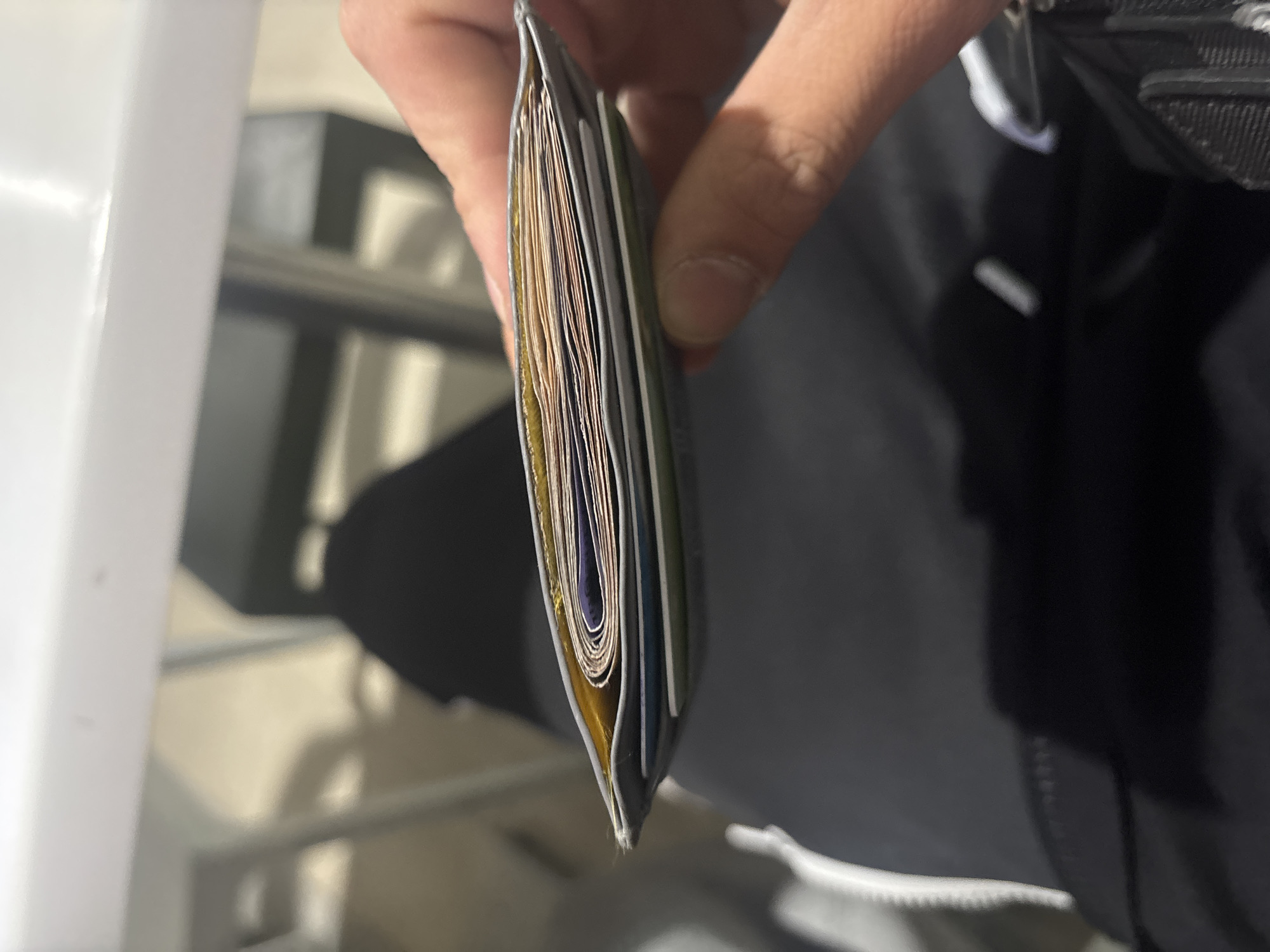 A person's hand holds a heavily stuffed, gray wallet or cardholder, presented vertically in the center of the frame. The wallet's interior is visible, revealing numerous layers of cards, cash, or papers in various colors, including prominent yellow/gold, purple/blue, and many lighter, possibly white or cream-colored layers. The hand has a medium skin tone. On the right side of the image, dark fabric, likely belonging to the person's clothing or a bag, is visible. The background is out of focus, showing blurry light-colored surfaces on the left, possibly a table or wall, and indistinct darker structures in the middle. The lighting appears bright, suggesting daytime conditions, either natural light from a window or strong artificial indoor lighting. No visible text is decipherable. The image depicts a close-up focus on the wallet and hand, with the surrounding environment blurred.