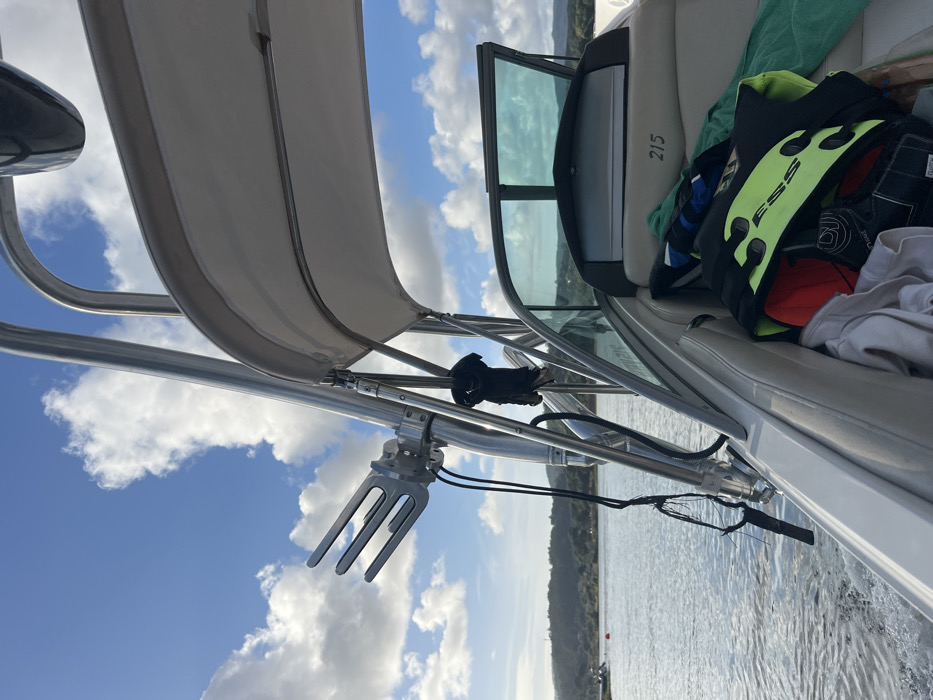The image depicts a forensic scene aboard a motorboat traversing a body of water, likely the Minho River, within or near Vila Nova de Cerveira, Portugal. The perspective is from within the boat, looking forward and to the port side.
In the foreground, a padded, white upholstered boat seat is visible, marked with the number "215" on its side. On this seat, various items are stored: a black and neon yellow personal flotation device or impact vest, a red baseball cap, white folded textiles (possibly towels or clothing), and a black wakeboarding glove.
Dominating the left side of the frame is a polished metal wakeboard tower supporting a white canvas bimini top, which provides shade. Attached to the tower is a silver-colored, three-pronged wakeboard rack, designed to hold a wakeboard. A black glove hangs from the tower, and black cables or ropes are visible, dangling from the tower structure. A black, cylindrical object, likely a speaker or light, is also mounted on the tower.
To the right, a tinted boat windshield with a walk-through opening is visible, suggesting the boat's forward cabin or bow area.
Beyond the boat's immediate structure, a body of water shows ripples and a distinct wake trailing behind the vessel, indicating it is in motion. In the mid-ground on the water, a red buoy is present, marking a channel or hazard. The background features a distant, undulating green shoreline densely covered with vegetation and hills, under a blue sky with scattered white cumulus clouds. No individuals are visible in the scene.