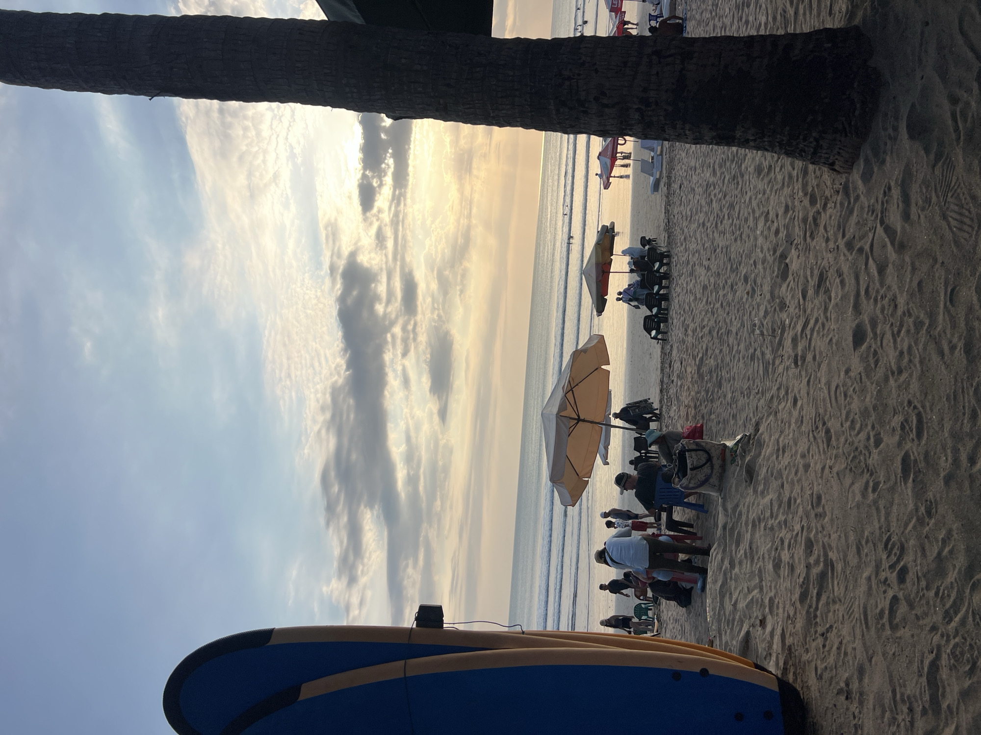 The image depicts a serene sunset scene at Kuta Beach in Bali, Indonesia. The foreground is dominated by the side of a blue and yellow surfboard, angled towards the left. Beyond the surfboard, a sandy beach stretches towards the ocean. Several people are scattered across the sand, some sitting under umbrellas and others standing. Two umbrellas, one beige and one red and yellow, provide shade for groups of people. Beach chairs are visible, some occupied. The ocean is calm with gentle waves rolling in. In the distance, more people can be seen in the water. The sky is a canvas of soft clouds, with the sun setting behind them, casting a warm, golden light across the scene. A tall palm tree trunk is prominently positioned on the right side of the image, partially obscuring the view of the sky and the background. The overall atmosphere is one of relaxation and leisure.